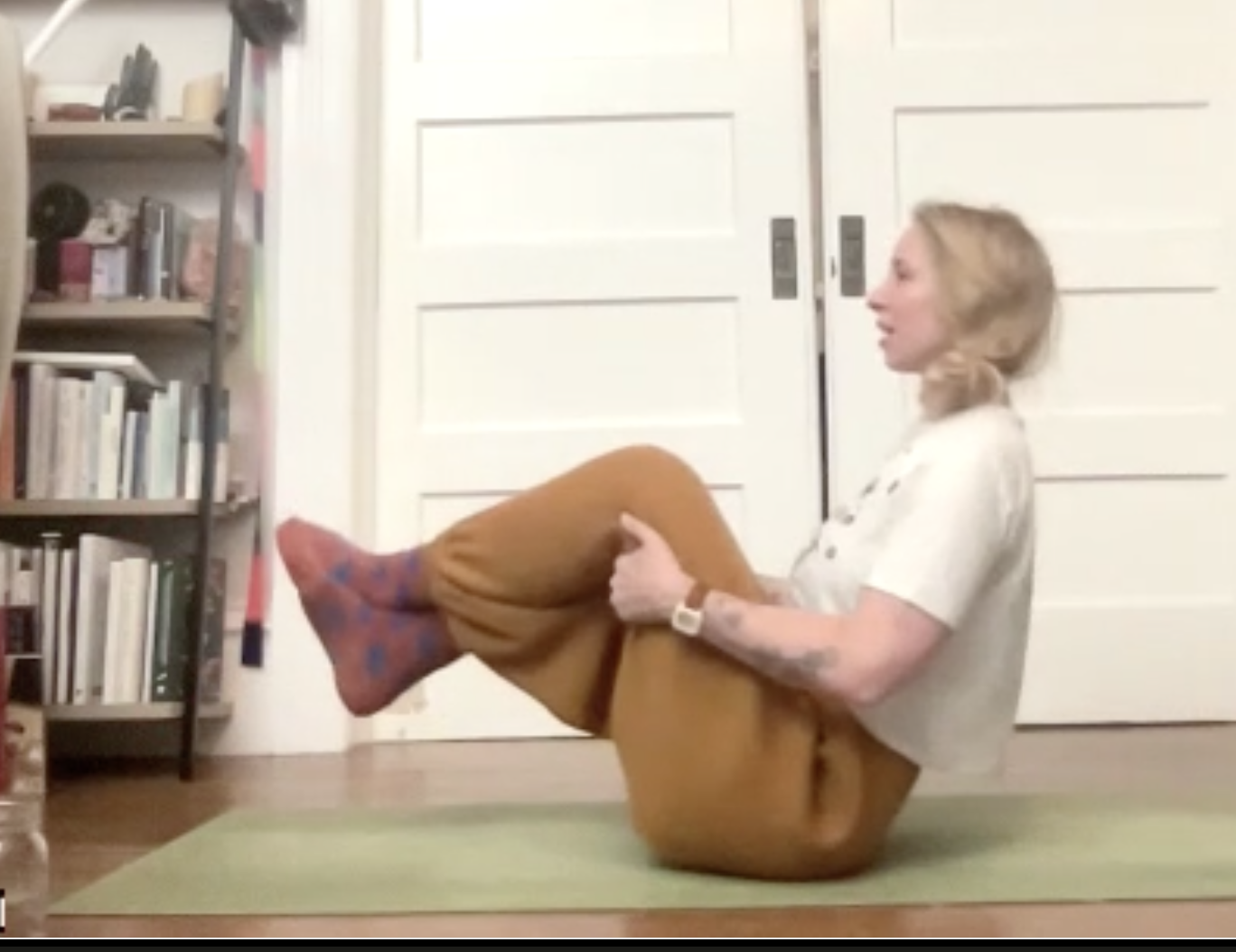 A woman practicing yoga on a green mat indoors, with bookshelves on the left and a white door behind her.