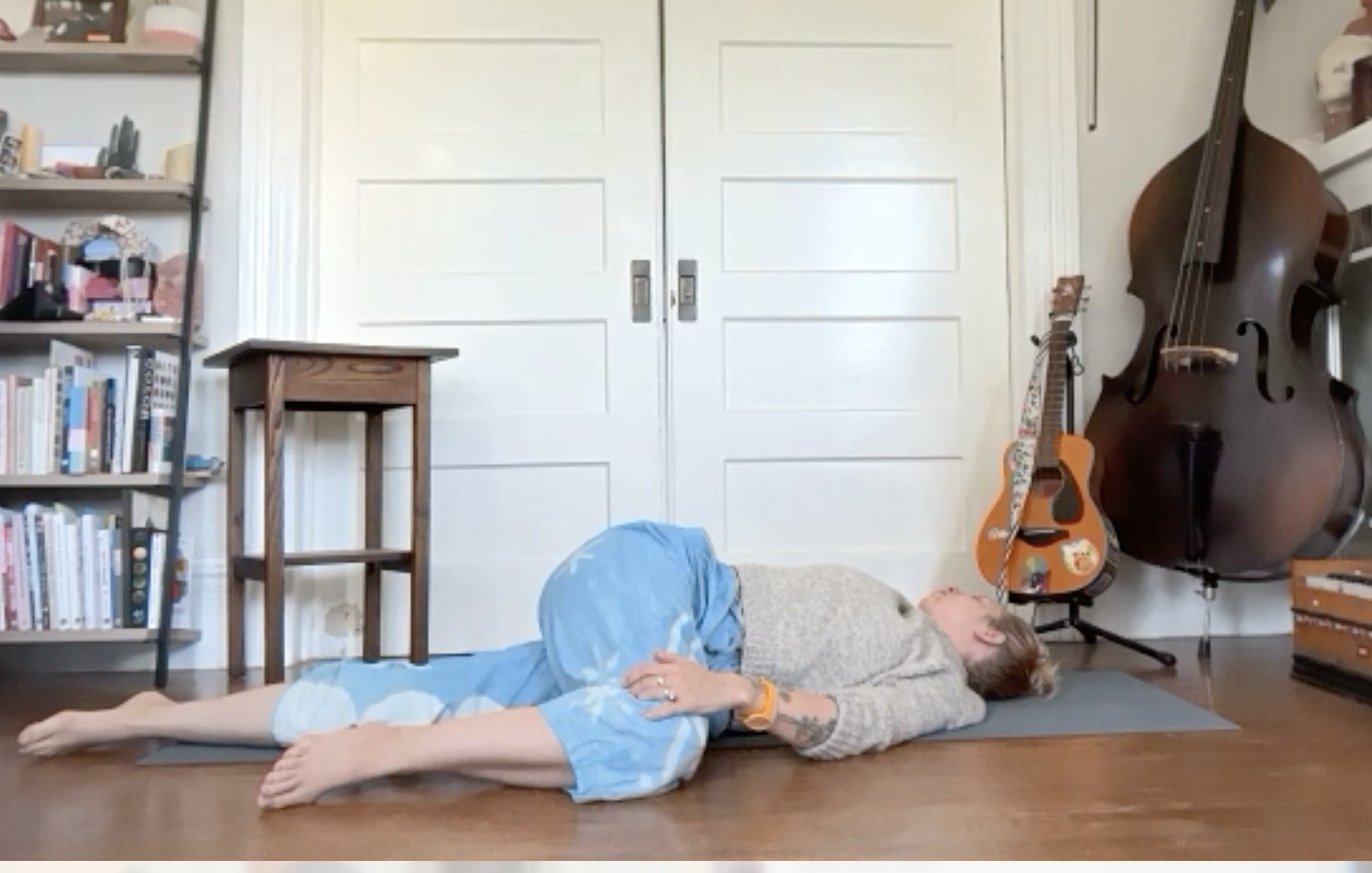 Person lying on yoga mat in a room with a bookshelf, a wooden stool, guitars, and a large cello in the background.