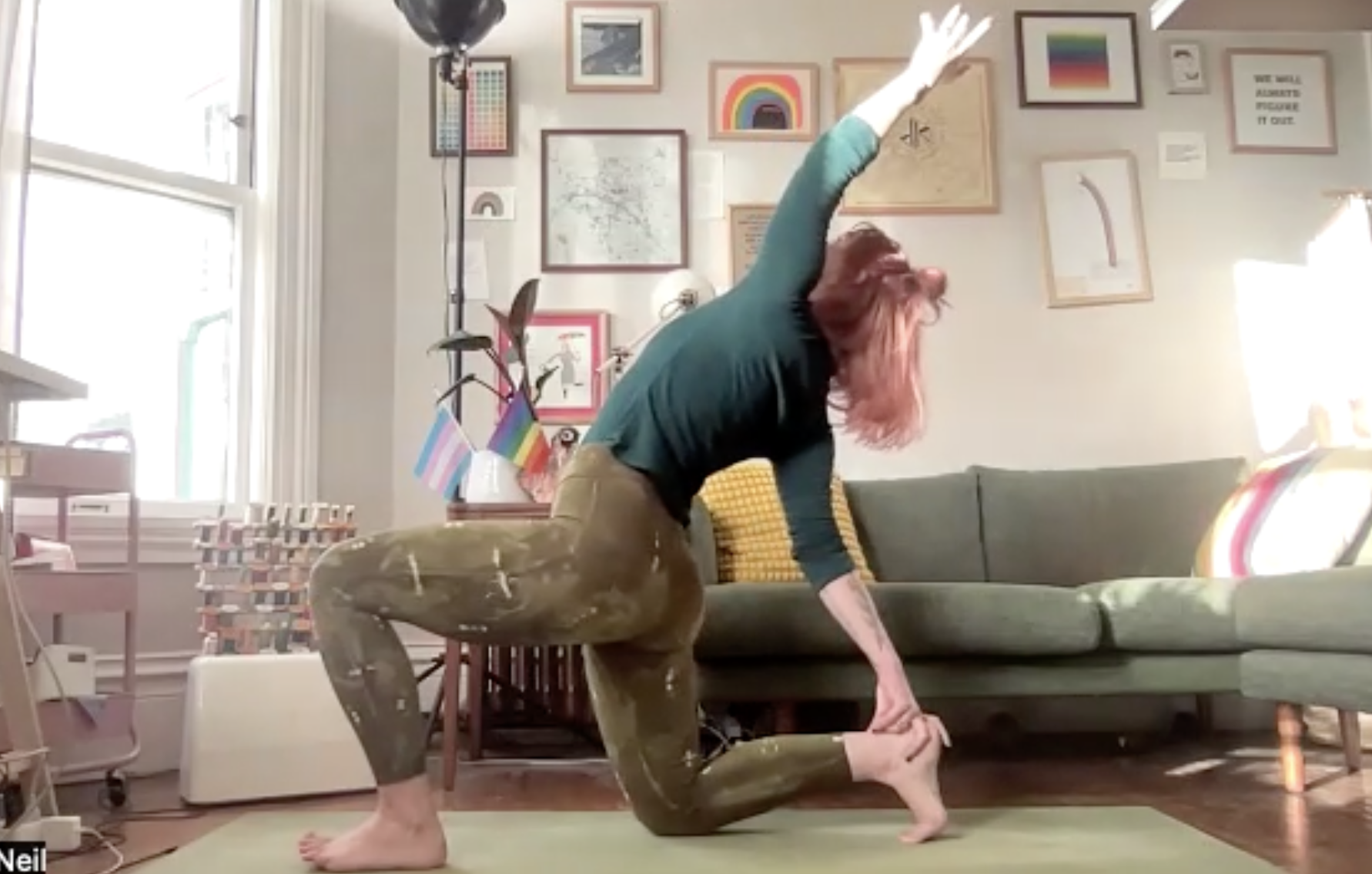 A woman with red hair practicing yoga in her living room, in a lizard pose with one hand on the floor and the other reaching upward. The room has framed artwork, a rainbow-themed print, LGBTQ+ flags on a table, a couch with yellow cushions, and brightly colored pillows. There is sunlight coming through a large window.