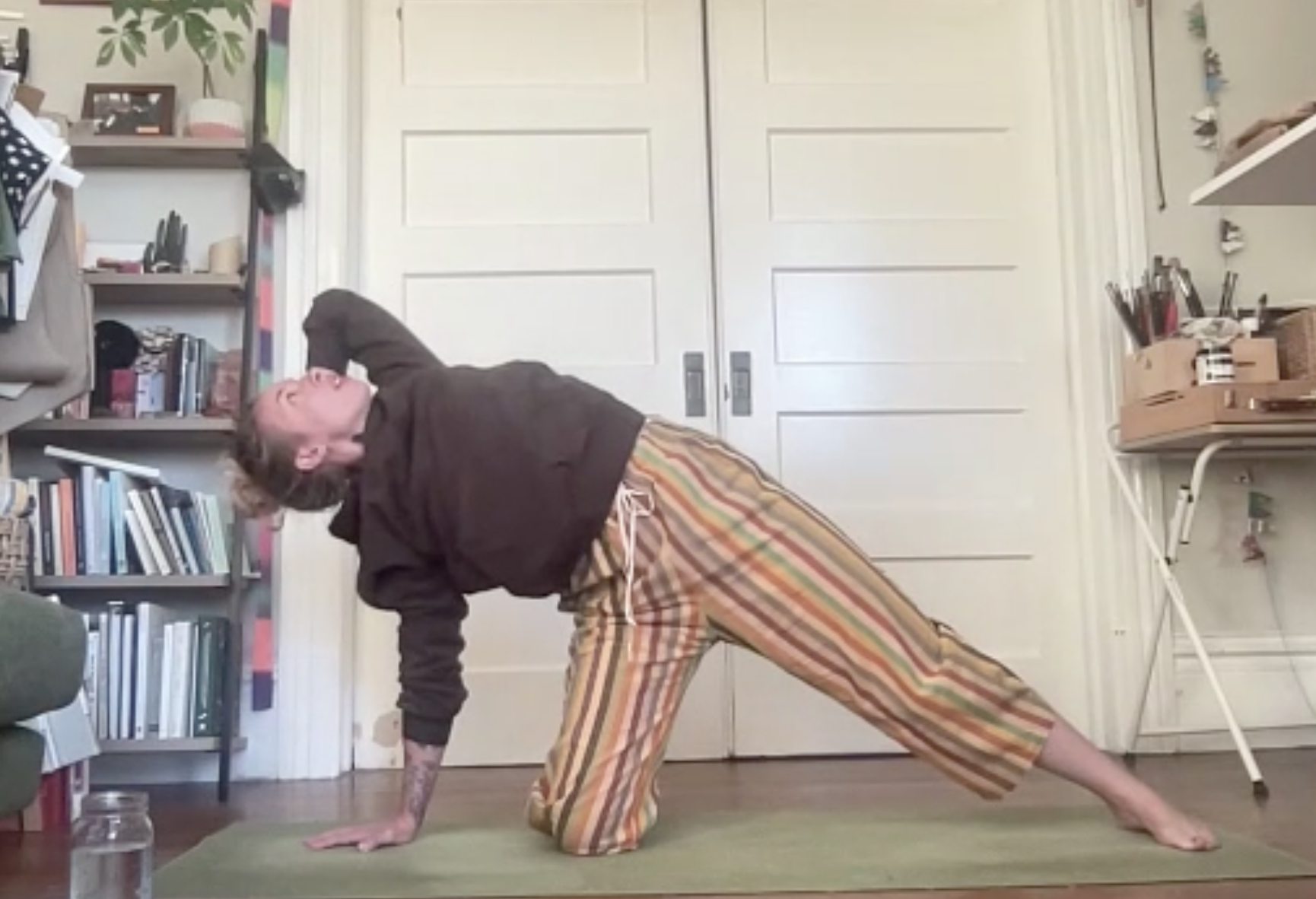 Person practicing yoga or stretching indoors, on a green mat, wearing striped pants and a black top, with bookshelves and shelves with art supplies in the background.