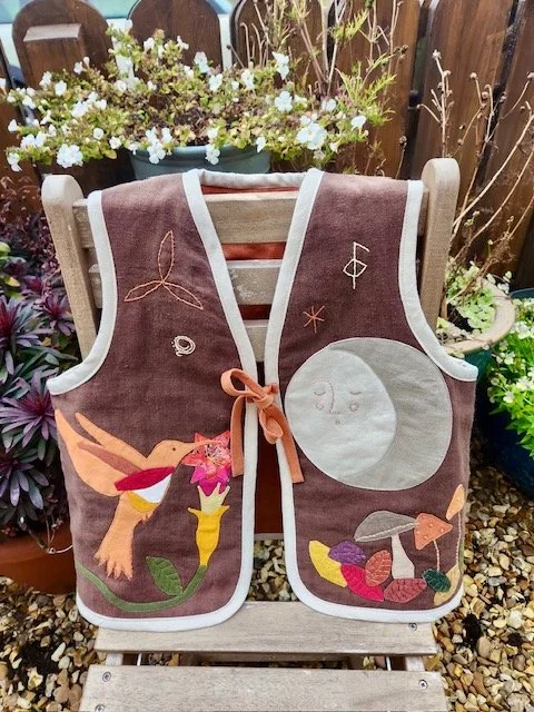Decorative quilted vest with embroidered and appliqué design, featuring a bird, moon, stars, and colorful yarn balls, hanging on a wooden chair outdoors with plants.