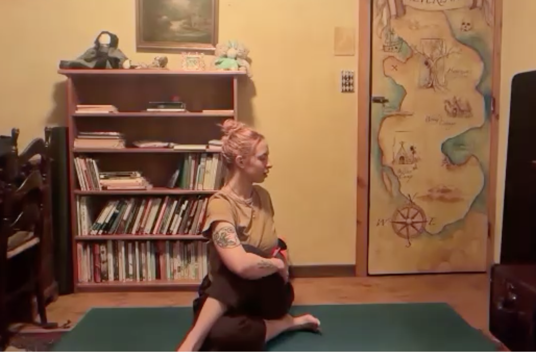 A woman with pink hair and tattoos practicing yoga on a green mat in a room with a bookshelf, a large map on the wall, and framed artwork.