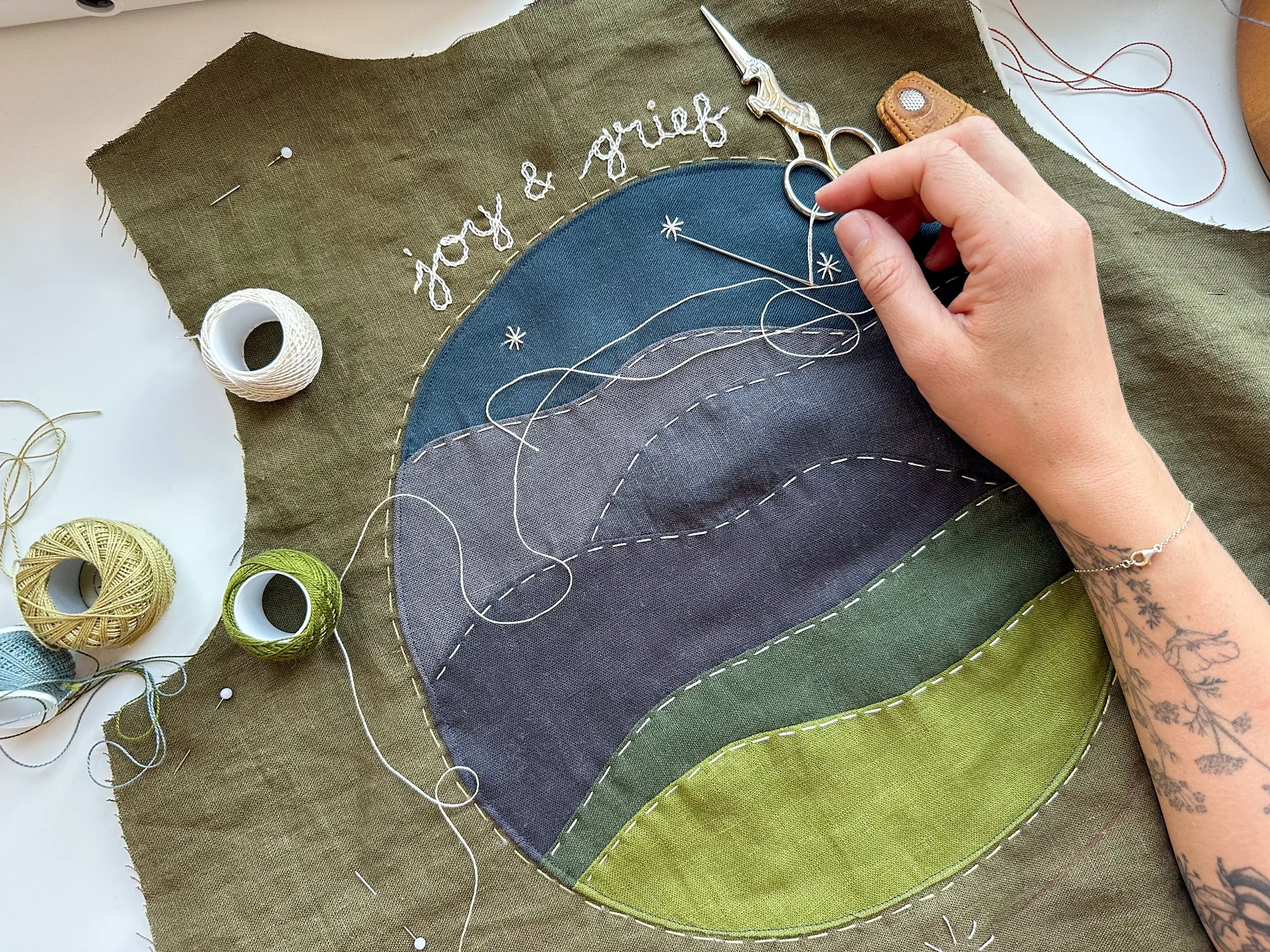 Close-up of a person stitching a landscape quilt with clouds and grassy hills, using colored fabric pieces, white thread, and sewing scissors. The quilt features embroidered text that reads 'joy & grief' and decorative star stitches.