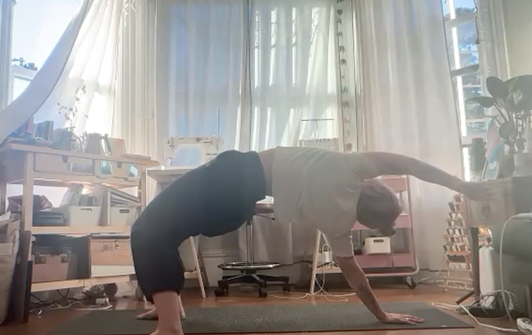 Person in a home office or living space practicing yoga or stretching in a downward dog position on a mat, with sunlight coming through large windows with white curtains. The room has bookshelves, a desk with a computer, plants, and various household items.