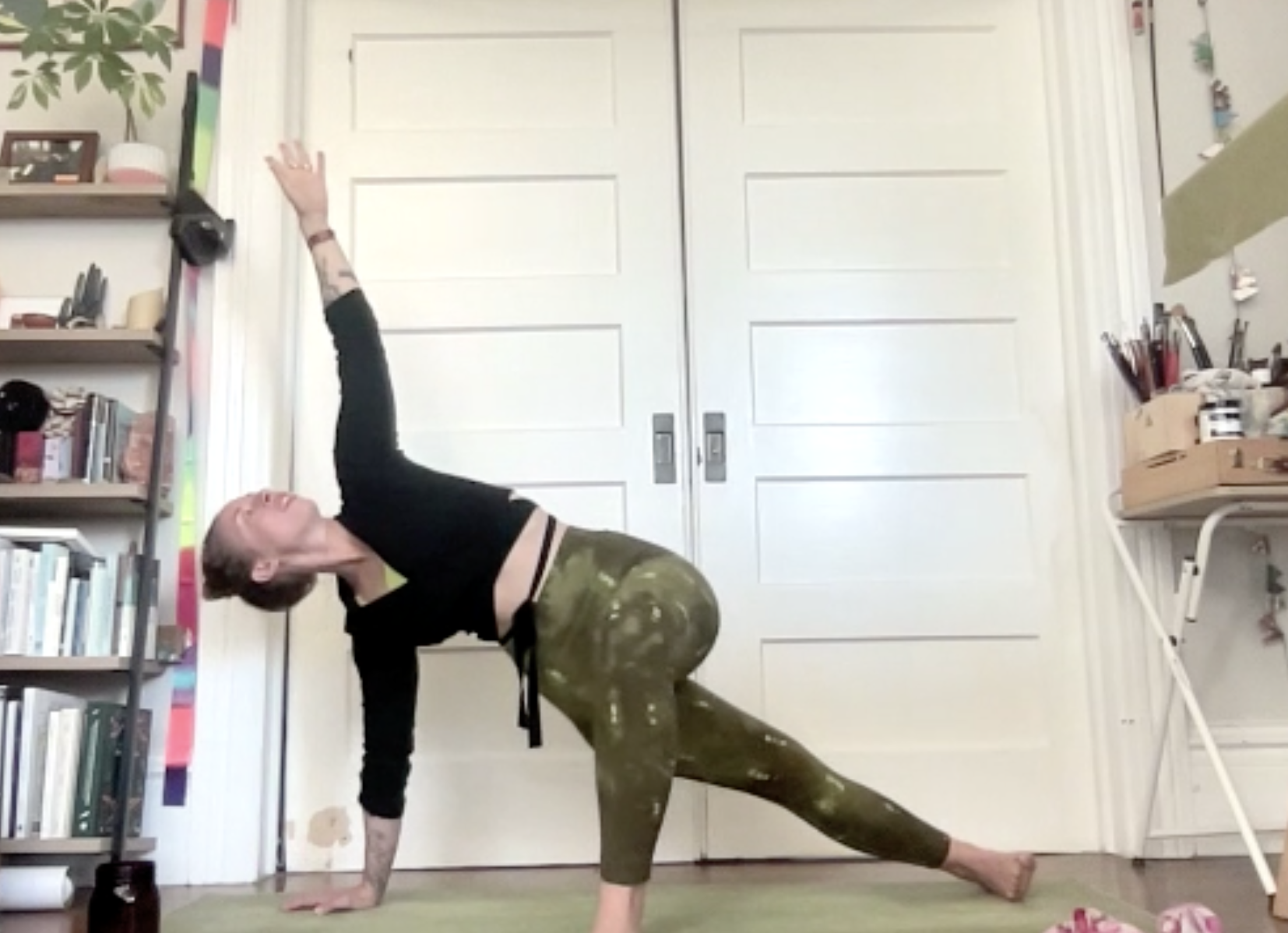 A woman practicing yoga indoors, performing a pose on a green yoga mat, with her left hand and right foot on the floor, and her right arm extended upward, in front of a white double door.