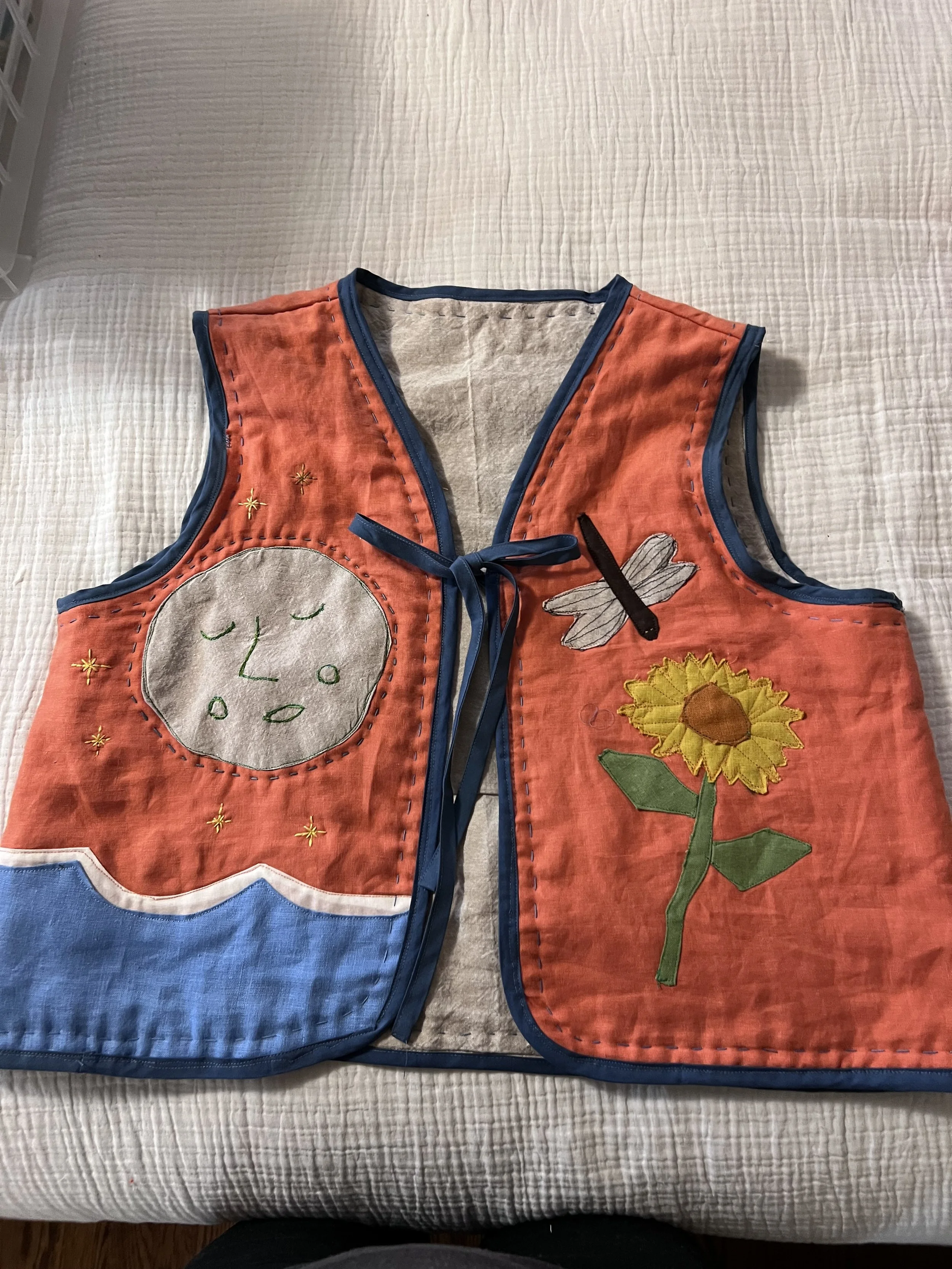 Child's sleeveless quilted vest with colorful appliqué design, featuring a moon with a face, a sunflower, and a dragonfly, on a white bedspread.