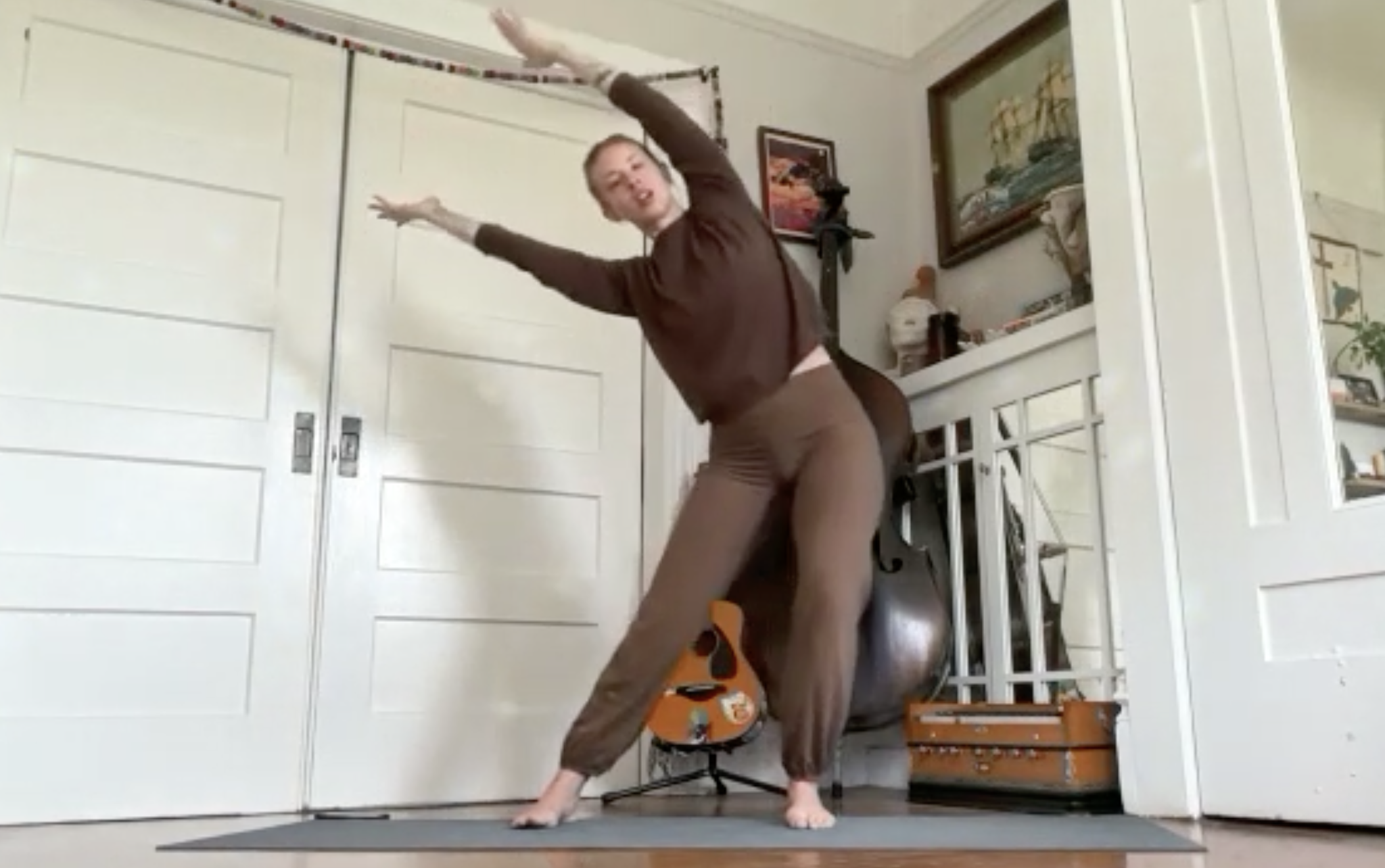 Person in a brown long-sleeve shirt and patterned shorts performing a yoga pose on a black mat inside a room with white walls, paintings, and wooden furniture.