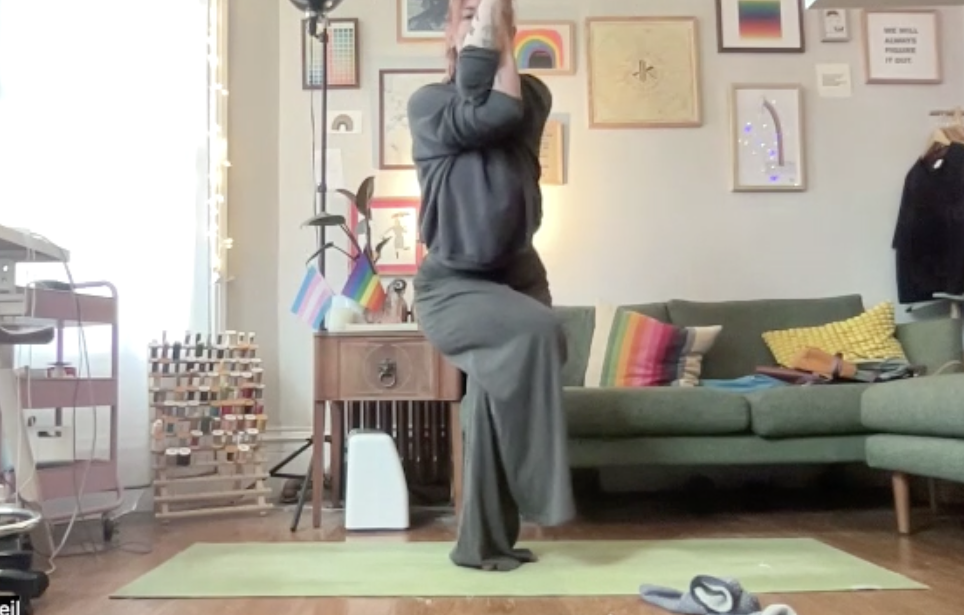 Person practicing yoga in a living room with art on the walls, a green couch with colorful pillows, and various household items.