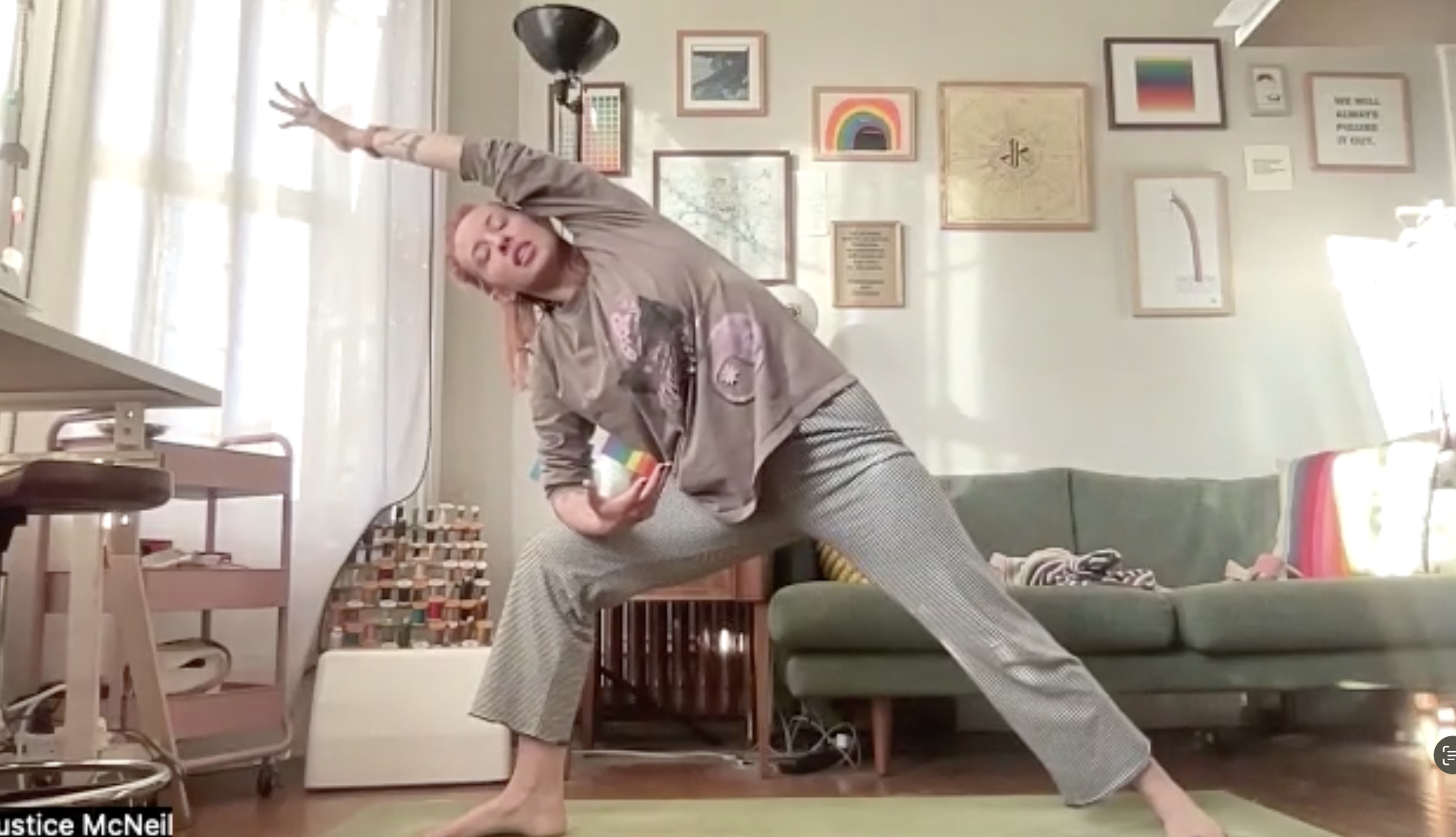 A woman practicing yoga in a living room, standing in a wide-legged stance with one arm extended upward and the other reaching down, wearing a gray sweatshirt and checkered pants, with art and photos on the wall behind her.
