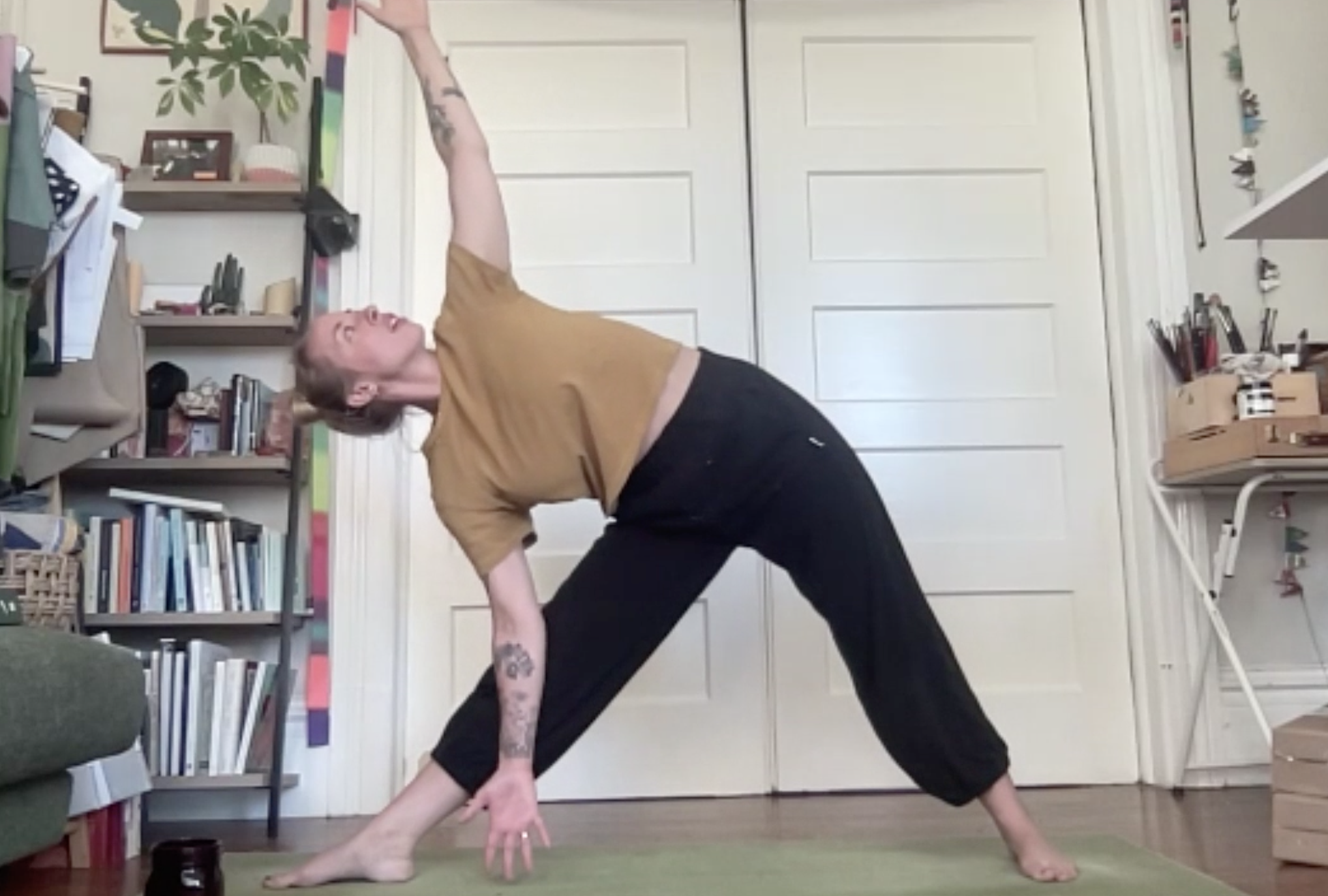 Person with tattoos performing a yoga pose in a room with bookshelves and closed white double doors.