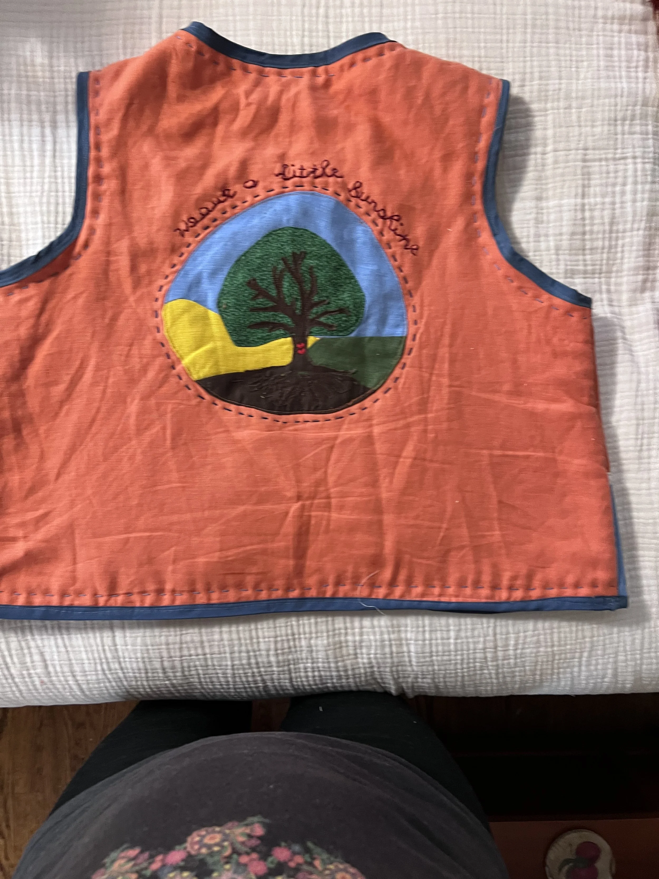 A pink fabric bib with embroidery of a tree and the words "Nature's Little Explorer" on a beige checkered surface.