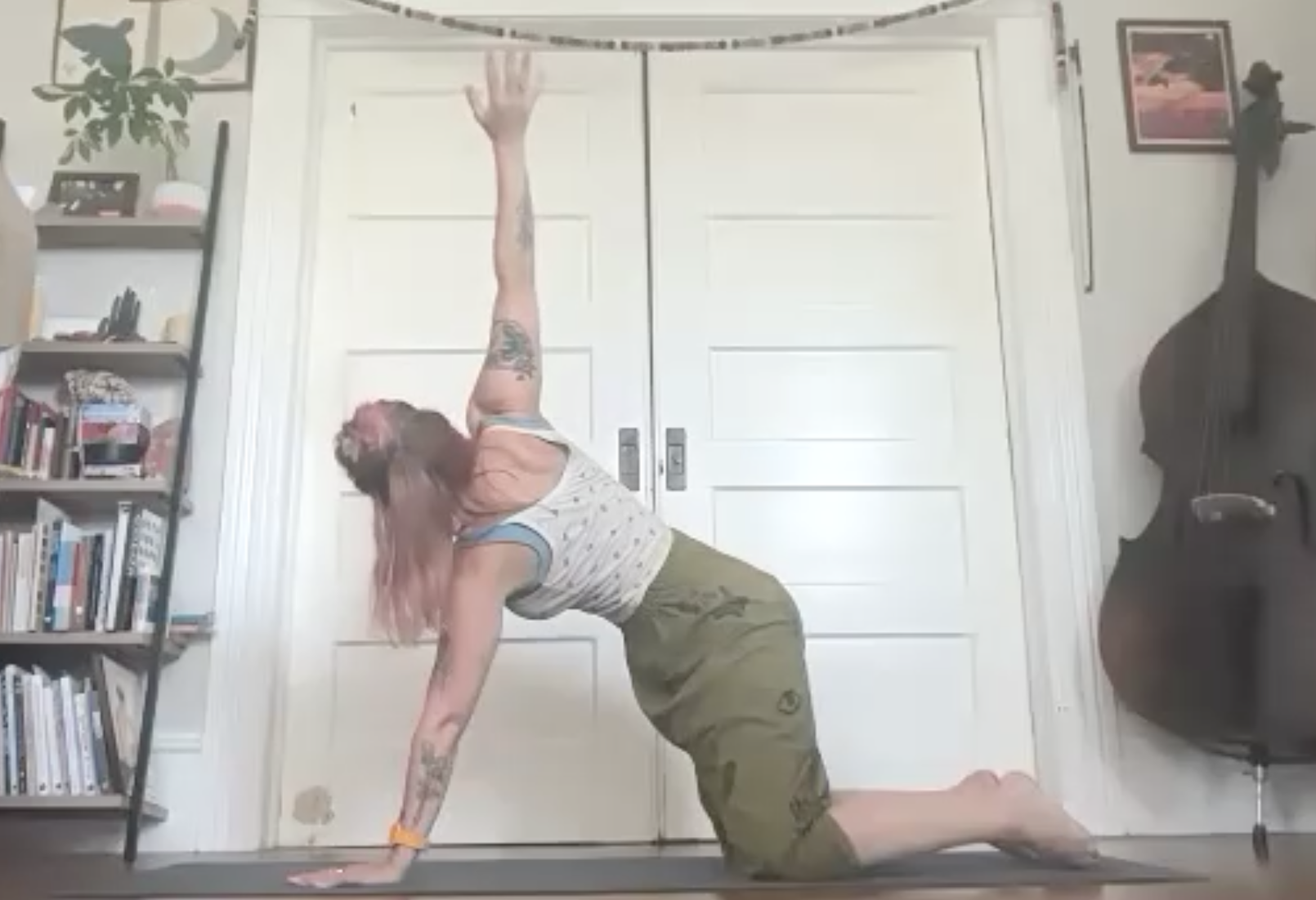 Person performing a yoga pose indoors, on a black mat, with a bookshelf and artwork visible in the background.