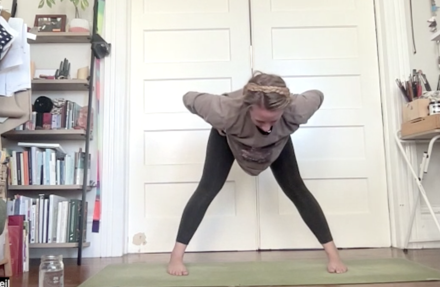 A woman practicing yoga in a room with bookshelves, a jar on the floor, and a closed white door. She is barefoot, wearing black leggings and a gray sweatshirt, and has her hair braided with a headband, while bending forward with her hands behind her back.
