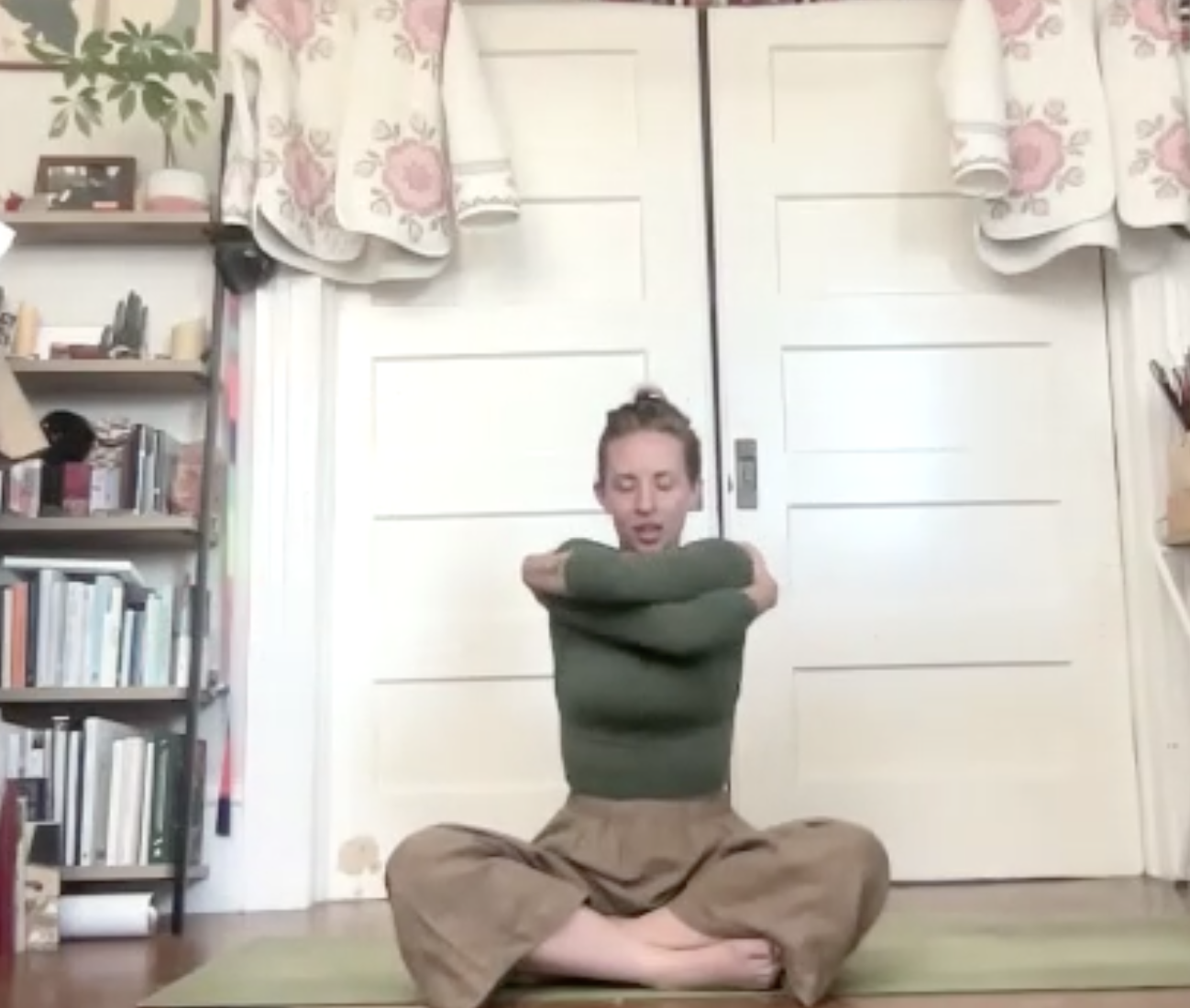 A woman practicing yoga or meditation in a room, sitting cross-legged on a mat with her arms crossed over her chest, eyes closed, and a calm expression.