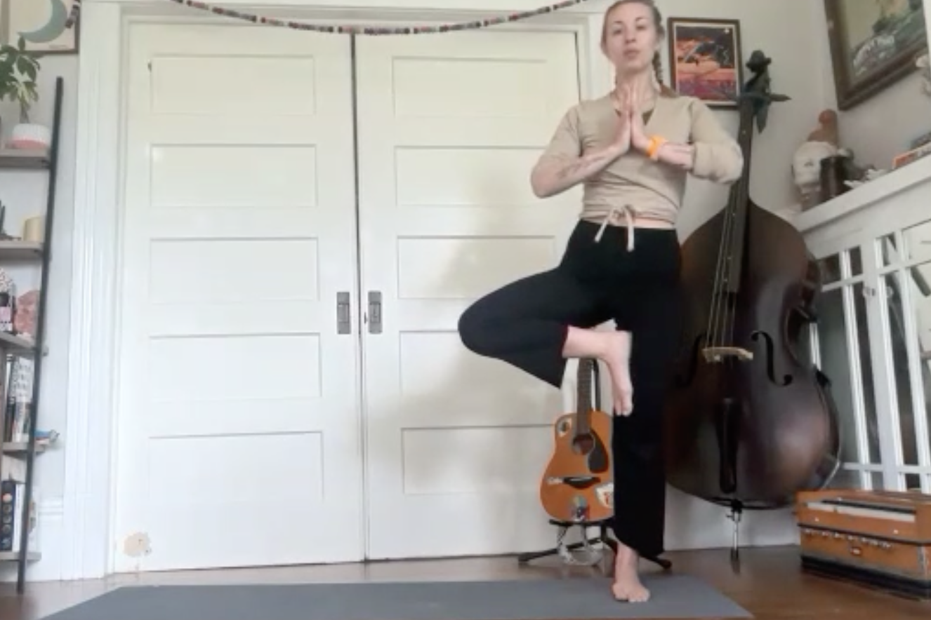 A woman practicing yoga indoors, standing on one leg with her hands in a prayer position, balancing on a yoga block, with a guitar and a double bass in the background.