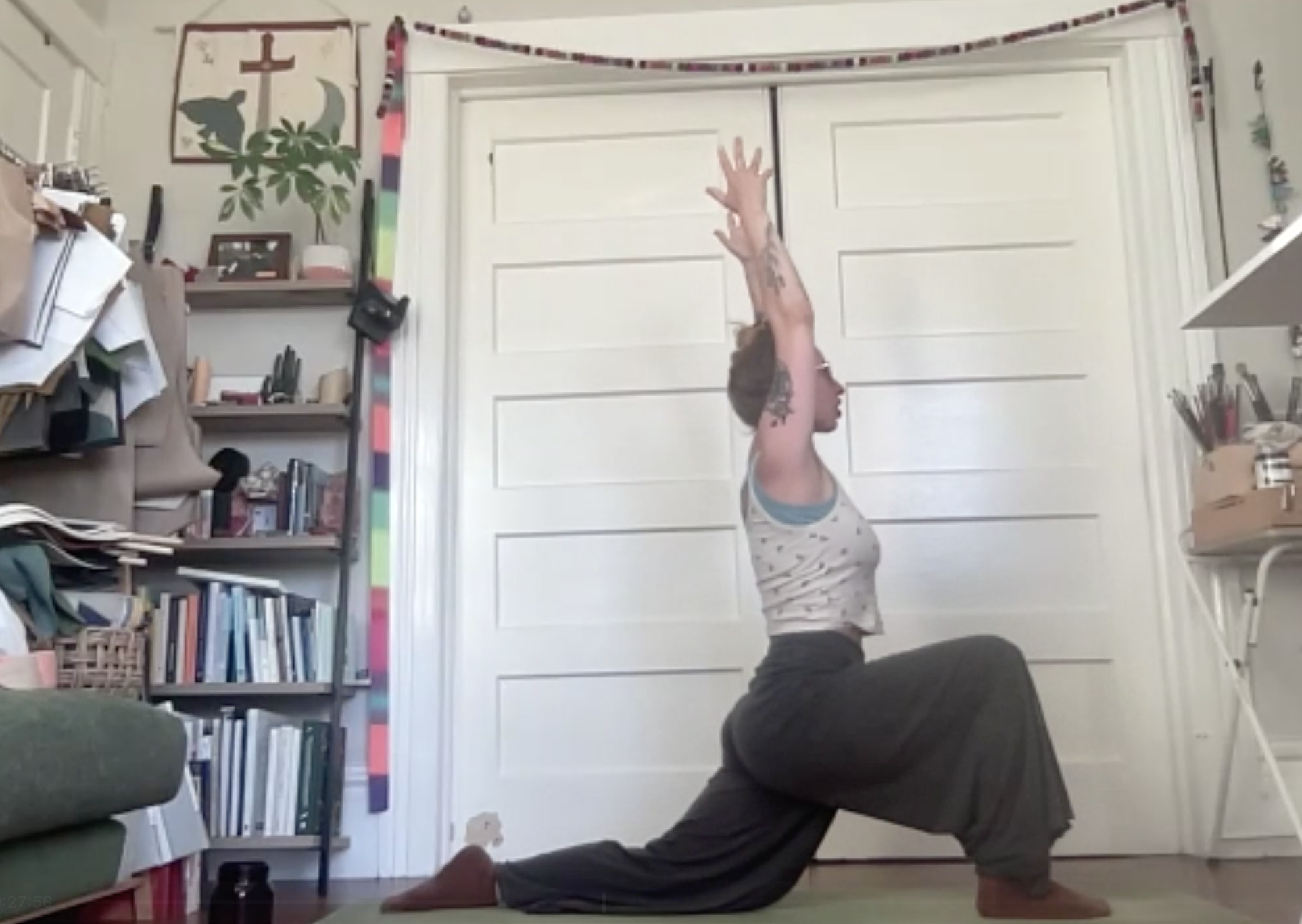 A person practicing yoga indoors, performing a downward facing dog pose with feet on the floor and hands reaching upward. The room has a bookshelf, a plant, and various decorations.