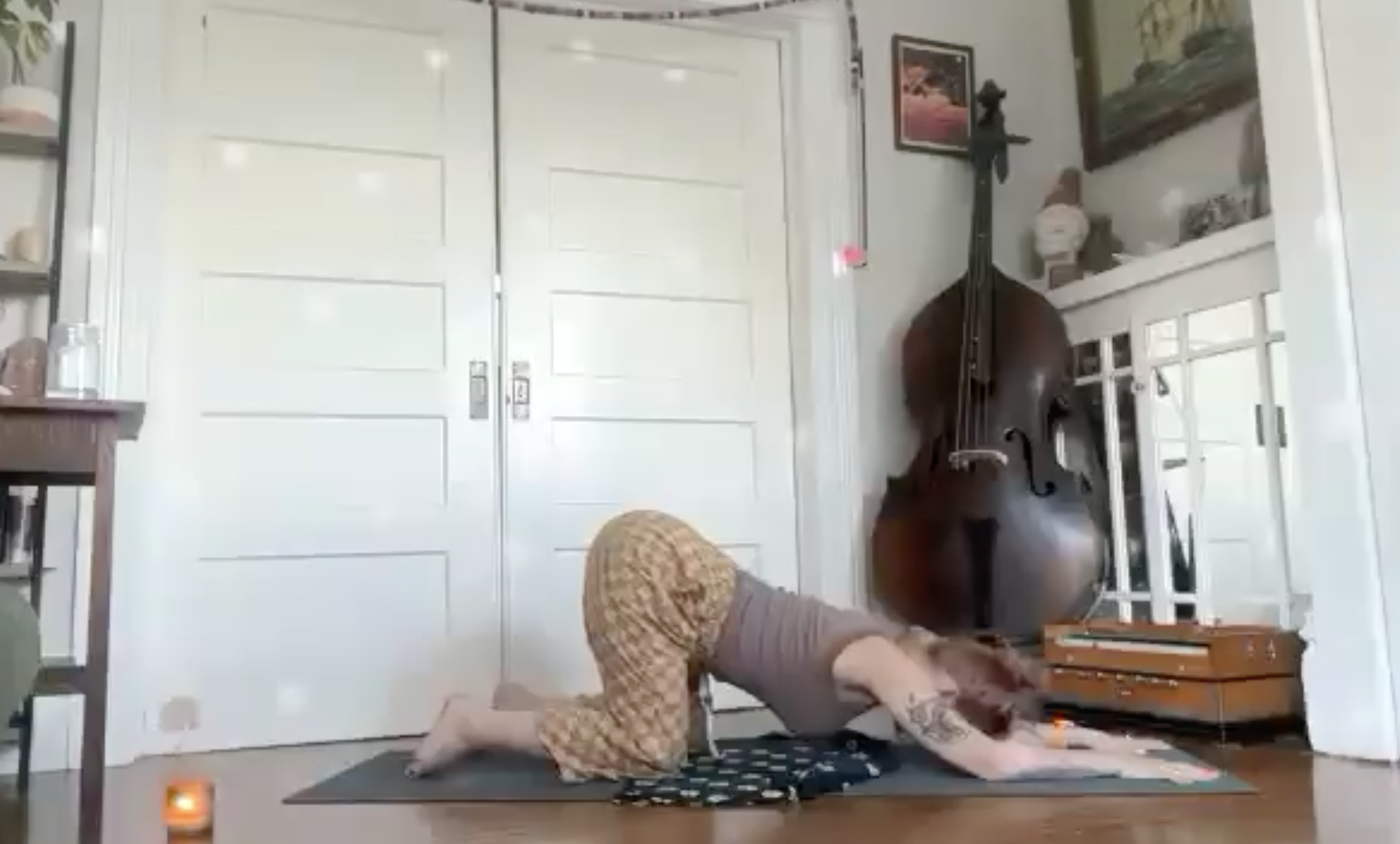 Person practicing yoga or stretching on a black mat in a room with white doors, a cello, and framed art on the walls.