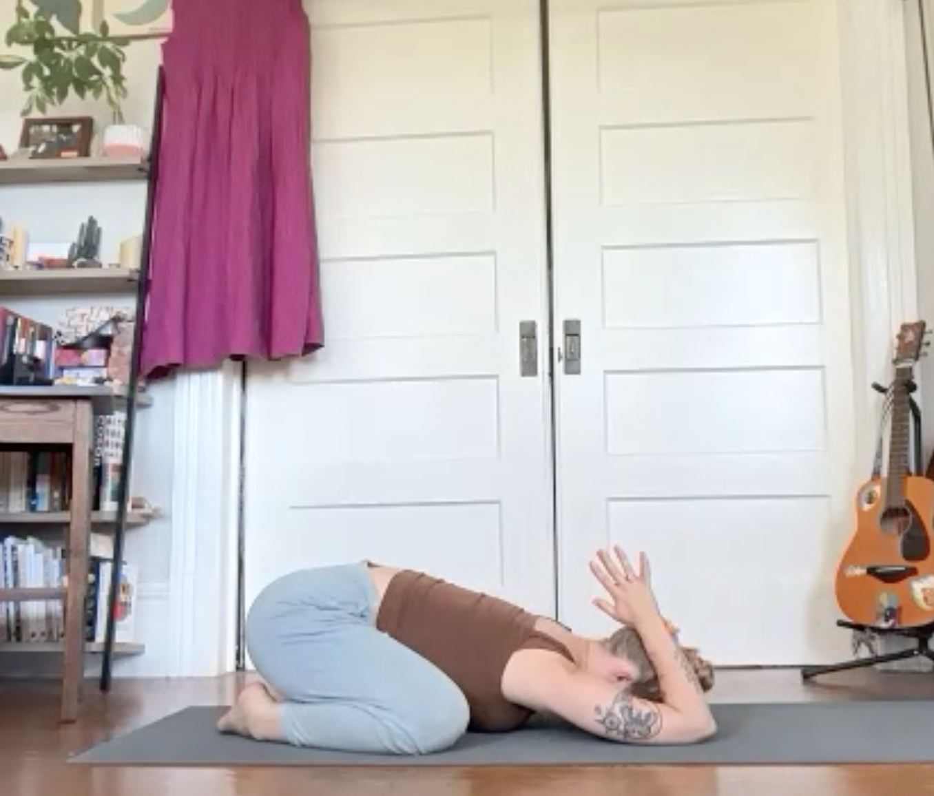 A person practicing yoga or meditation on a mat in a room with bookshelves, a guitar, and a closed closet door, with their hand raised in front of their face.