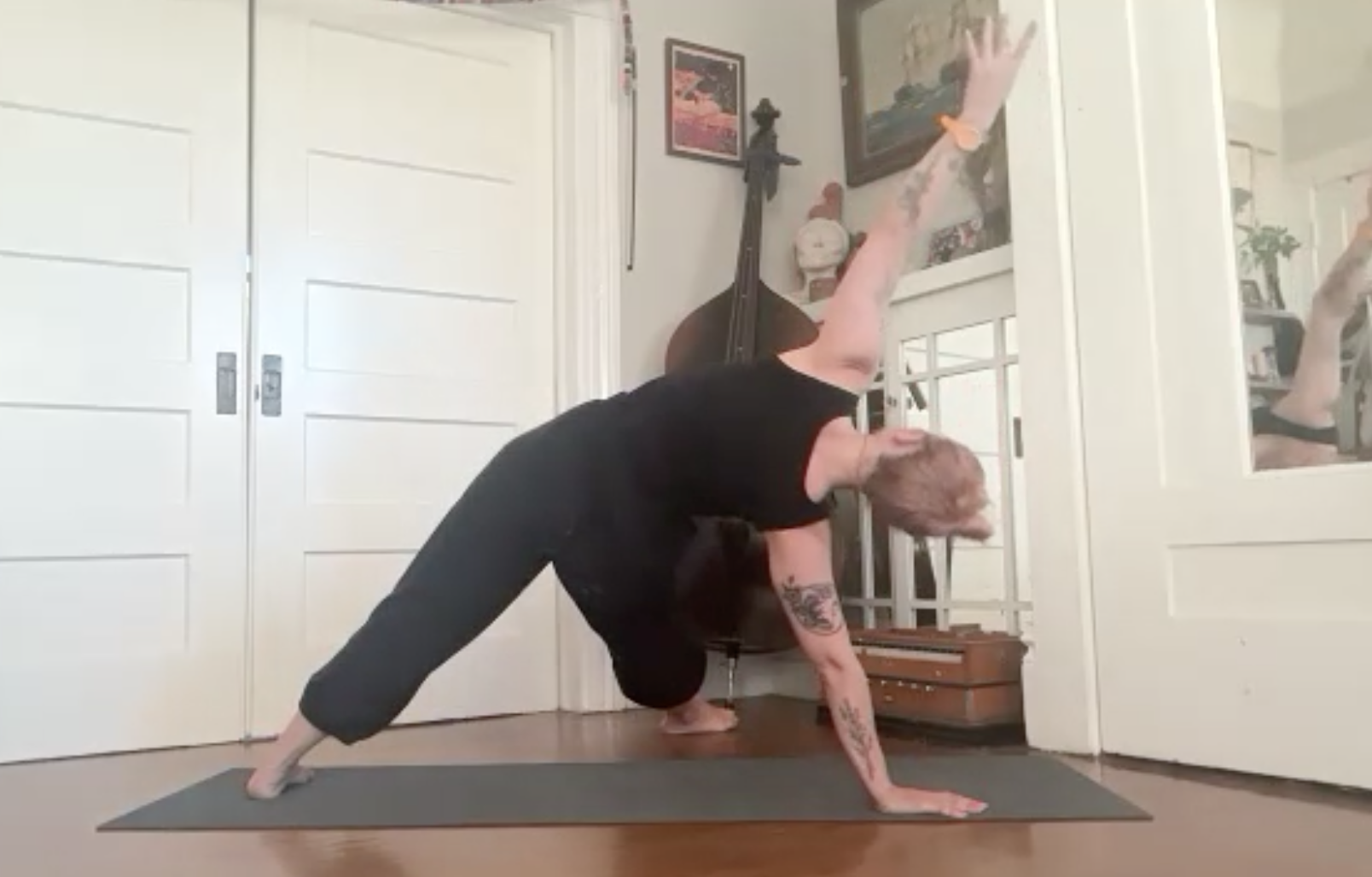 Person practicing yoga indoors on a mat, in a pose with one hand on the ground and the other hand extended upwards, with a guitar and framed artwork in the background.