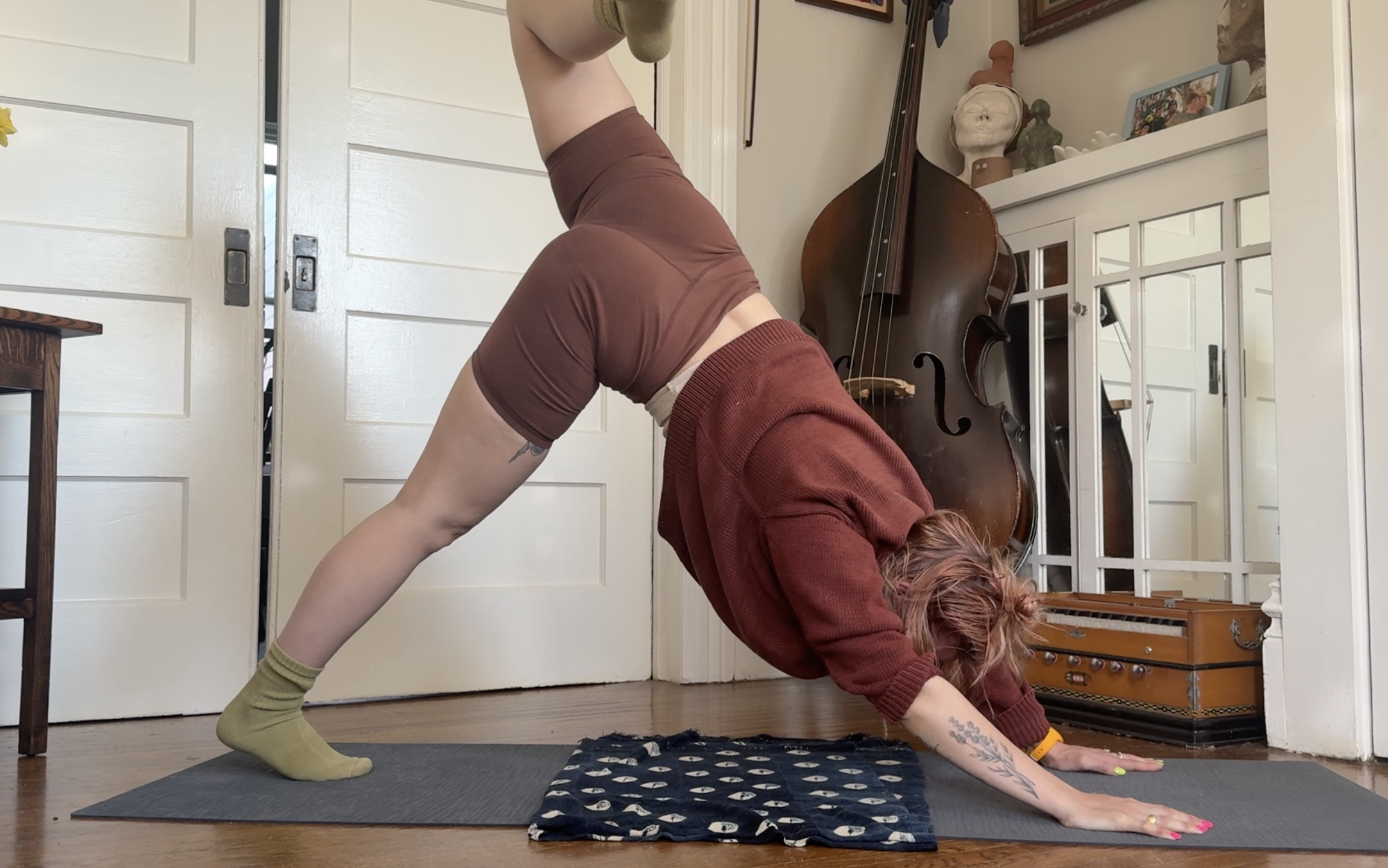 A person with pink hair and tattoos practicing yoga in a downward-facing dog pose on a black and white mat, with another person with short hair in a forward bend pose on the mat next to them, inside a room with a double bass, decorative statues, and a white cabinet.