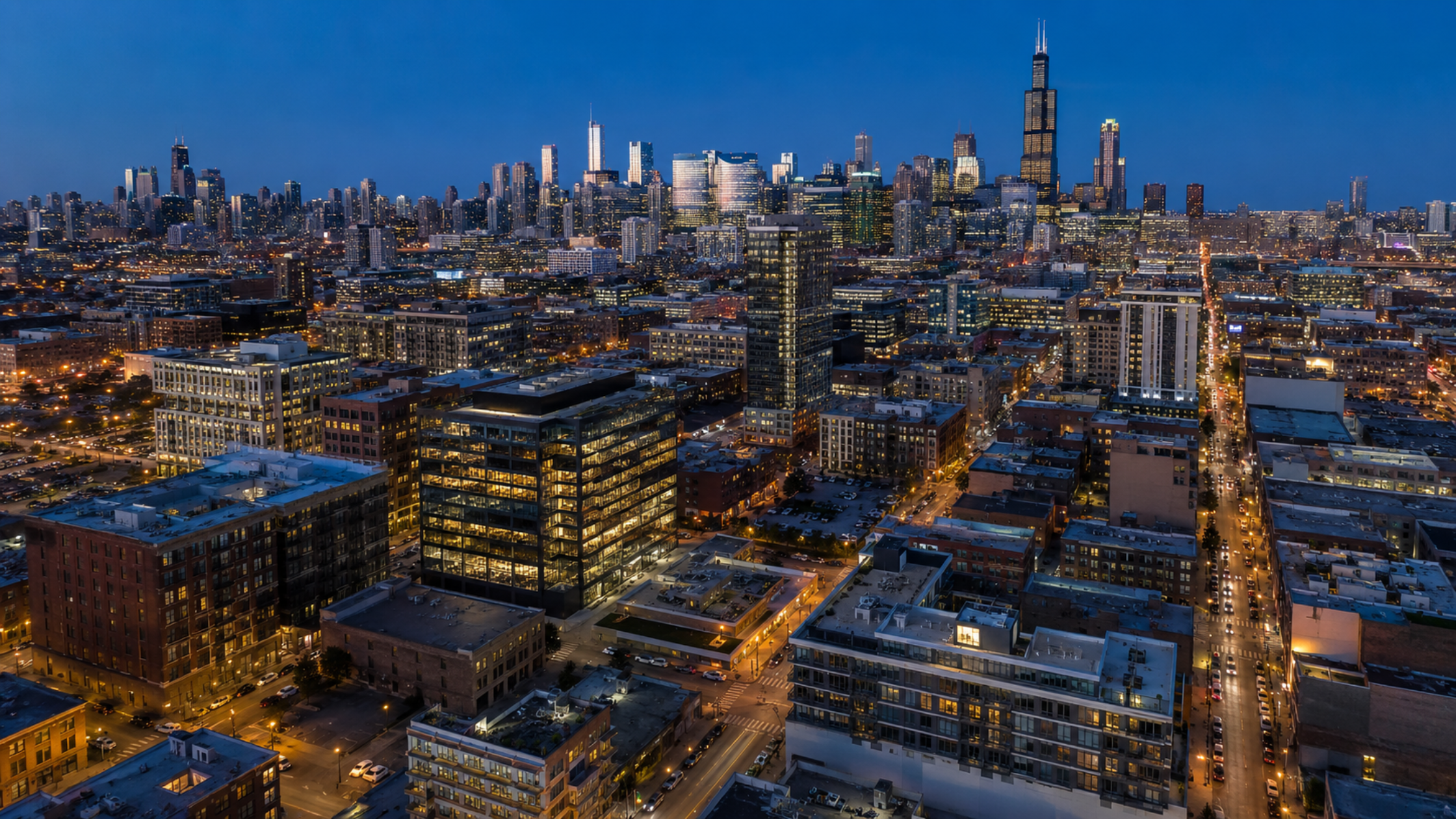 An aerial drone photo looking west over West Loop Chicago at dusk, with the city skyline glowing behind and Fulton Market streetlights illuminating the grid below.