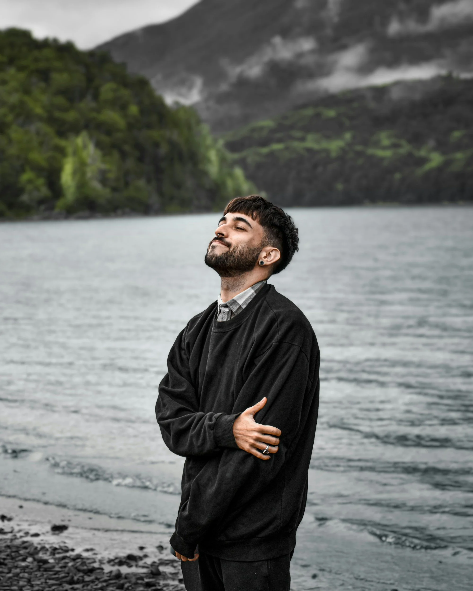 Person in black sweater standing by a lake with mountains in the background, looking serene.