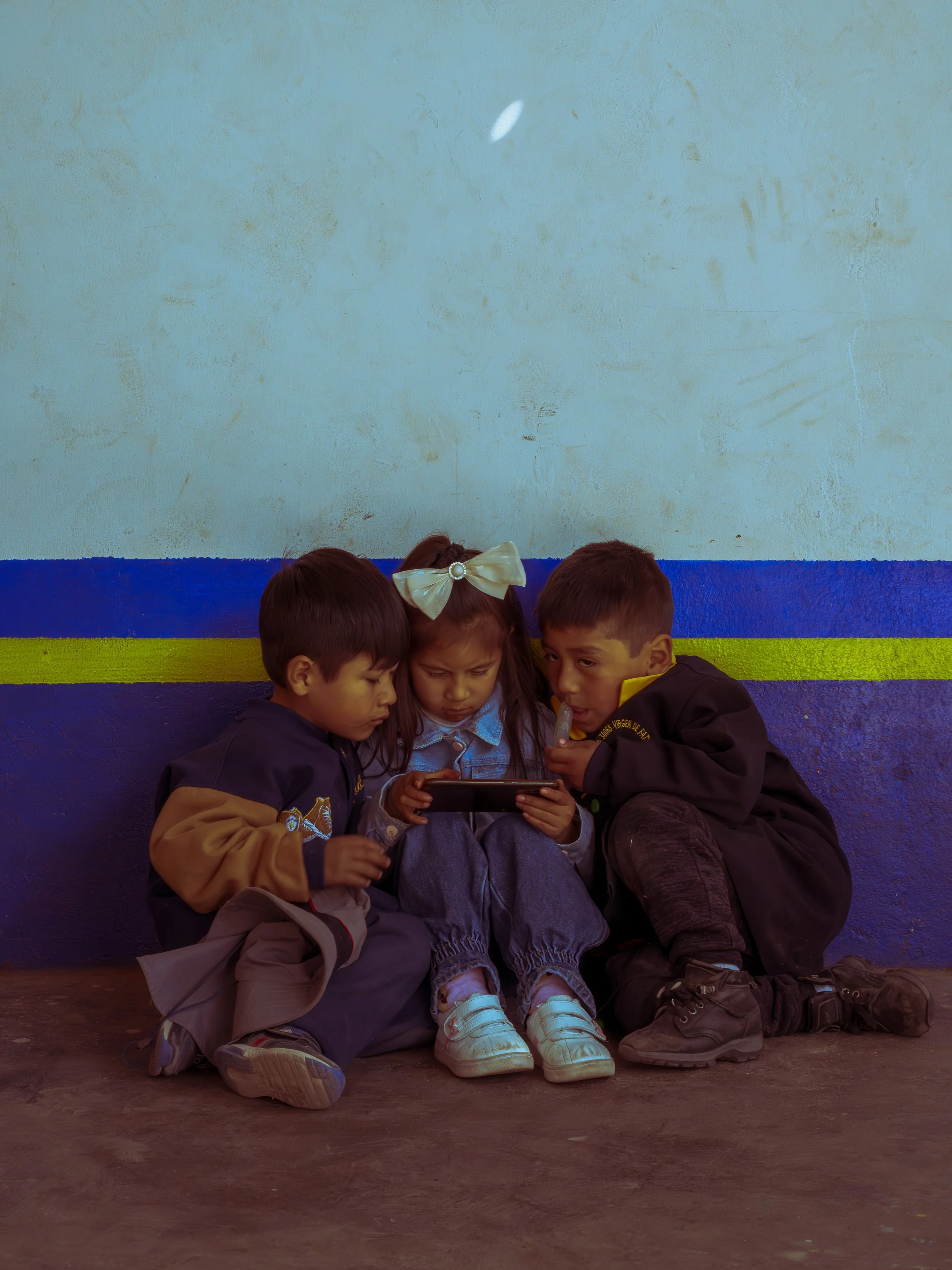 Three children sitting on the ground against a colorful wall, looking at a smartphone together.