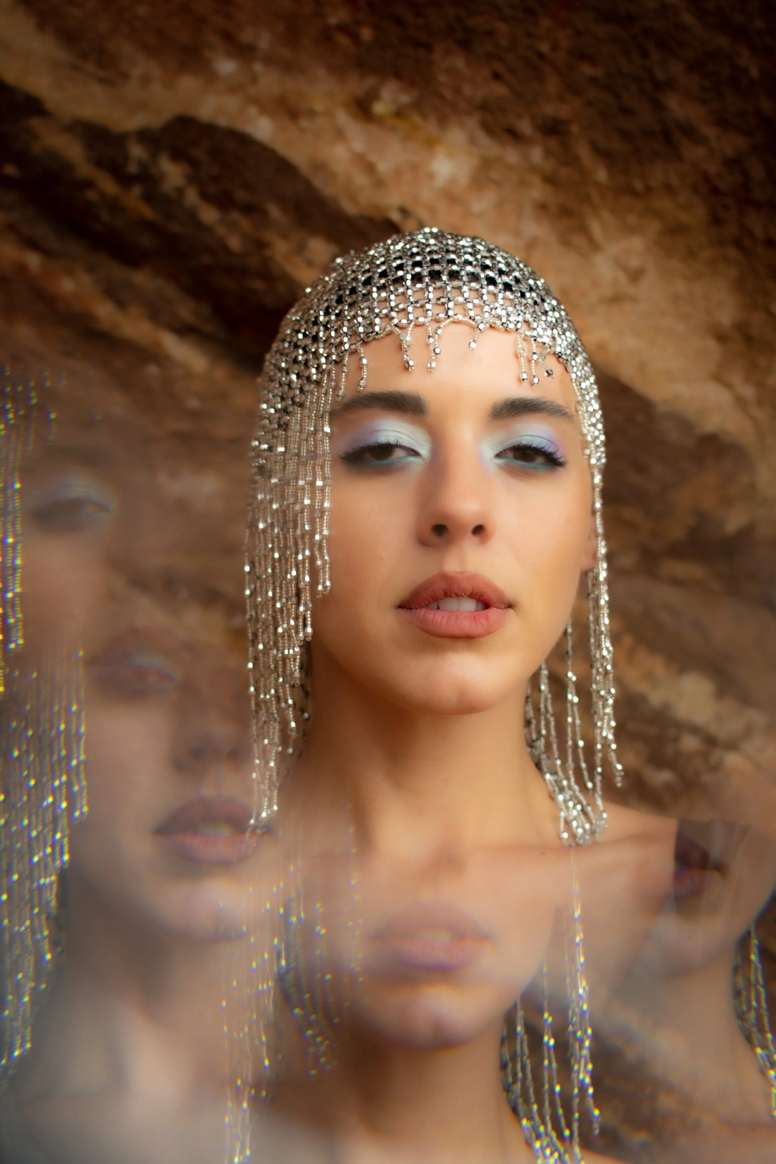 Close-up of a woman with silver jewelry and makeup, with a brown background and a blurred reflection.