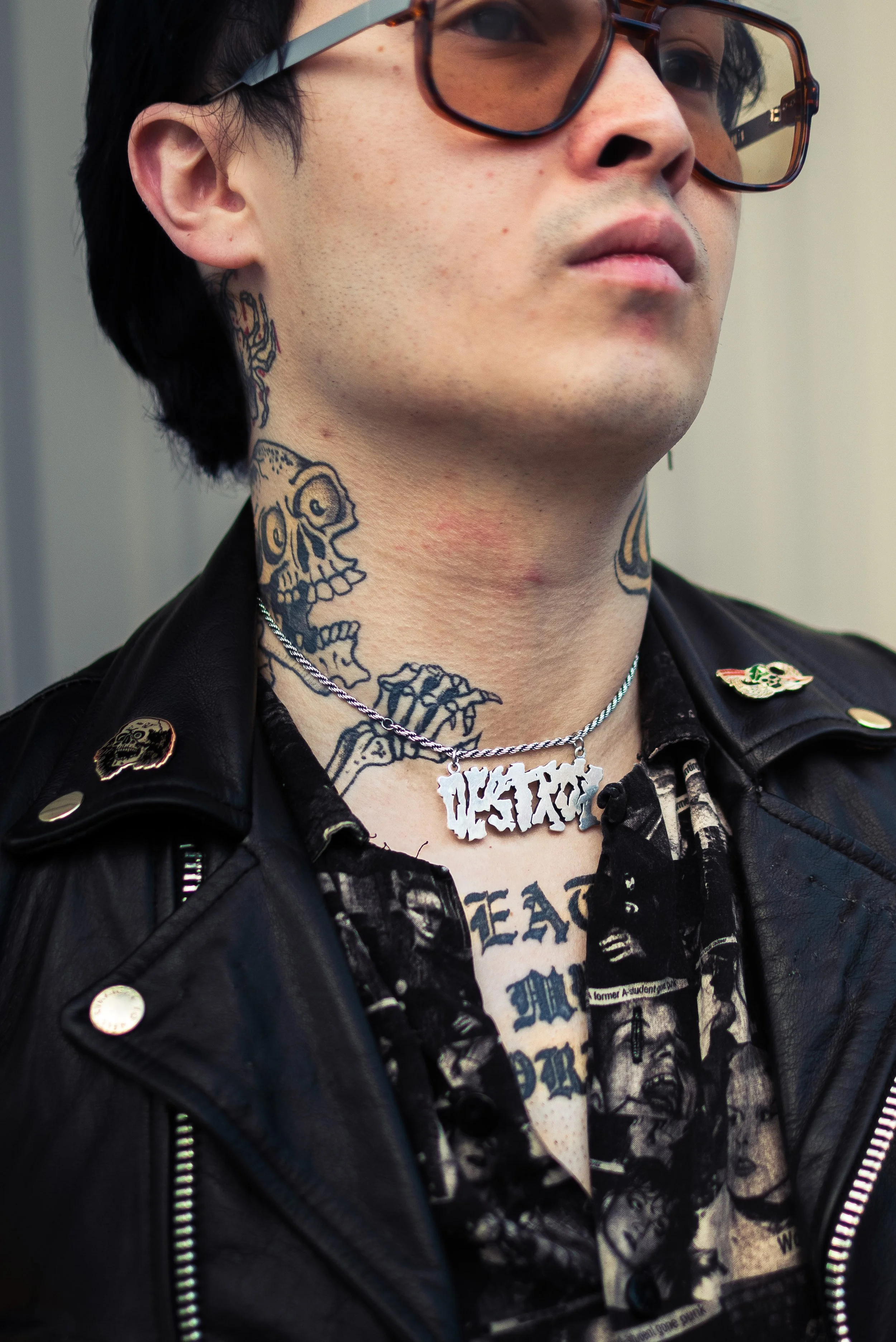 Close-up of a person with multiple tattoos on their neck and chest, wearing glasses, a leather jacket with patches, and a silver chain with a large, stylized pendant that appears to say 'PASTOR'.
