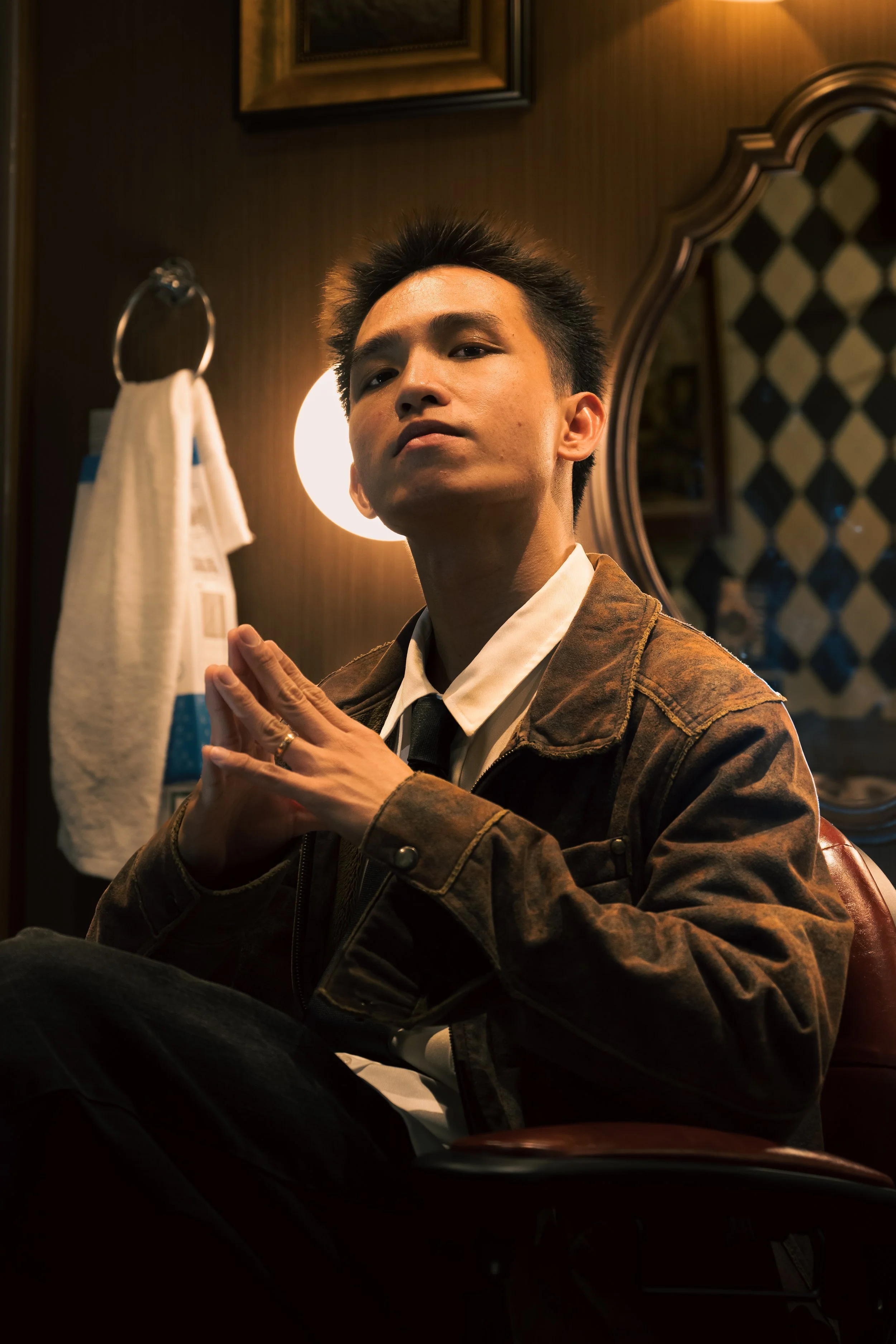 A young man with dark hair and a serious expression, wearing a brown jacket over a white shirt with a black tie, sitting indoors in front of a mirror, with warm lighting and patterned wallpaper in the background.