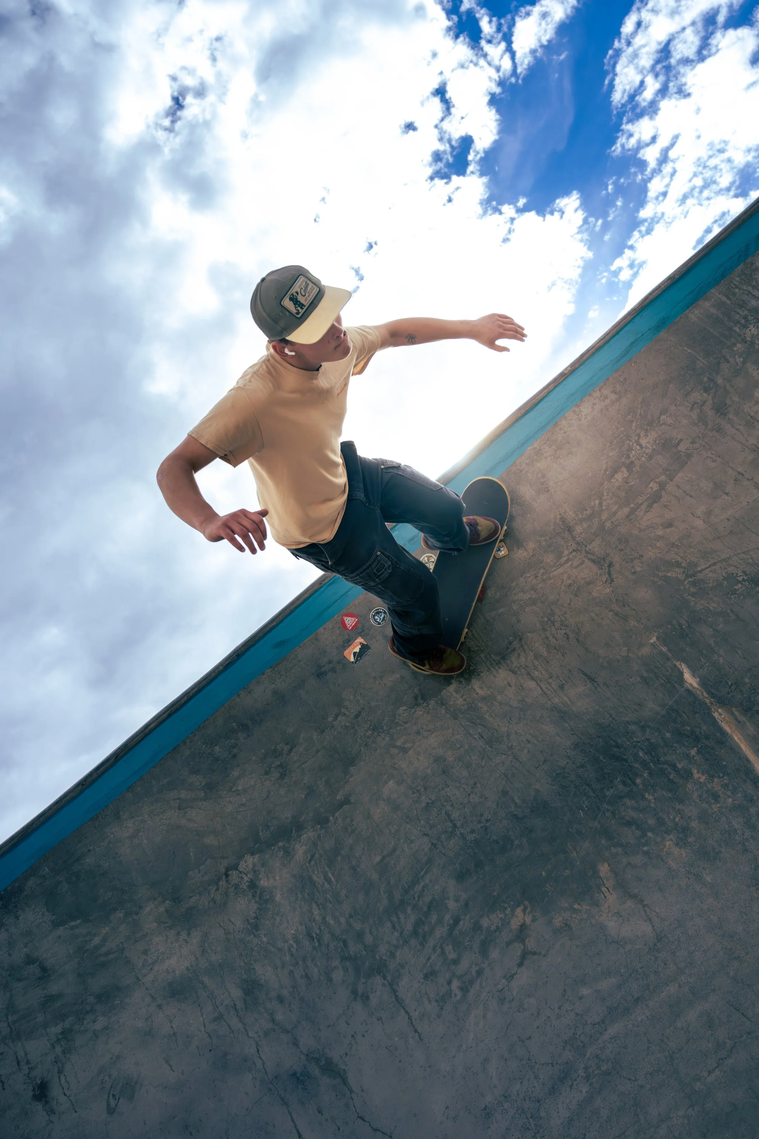 A person skateboarding outdoors on a concrete surface, wearing a baseball cap, beige t-shirt, and jeans, with a blue sky and clouds in the background.