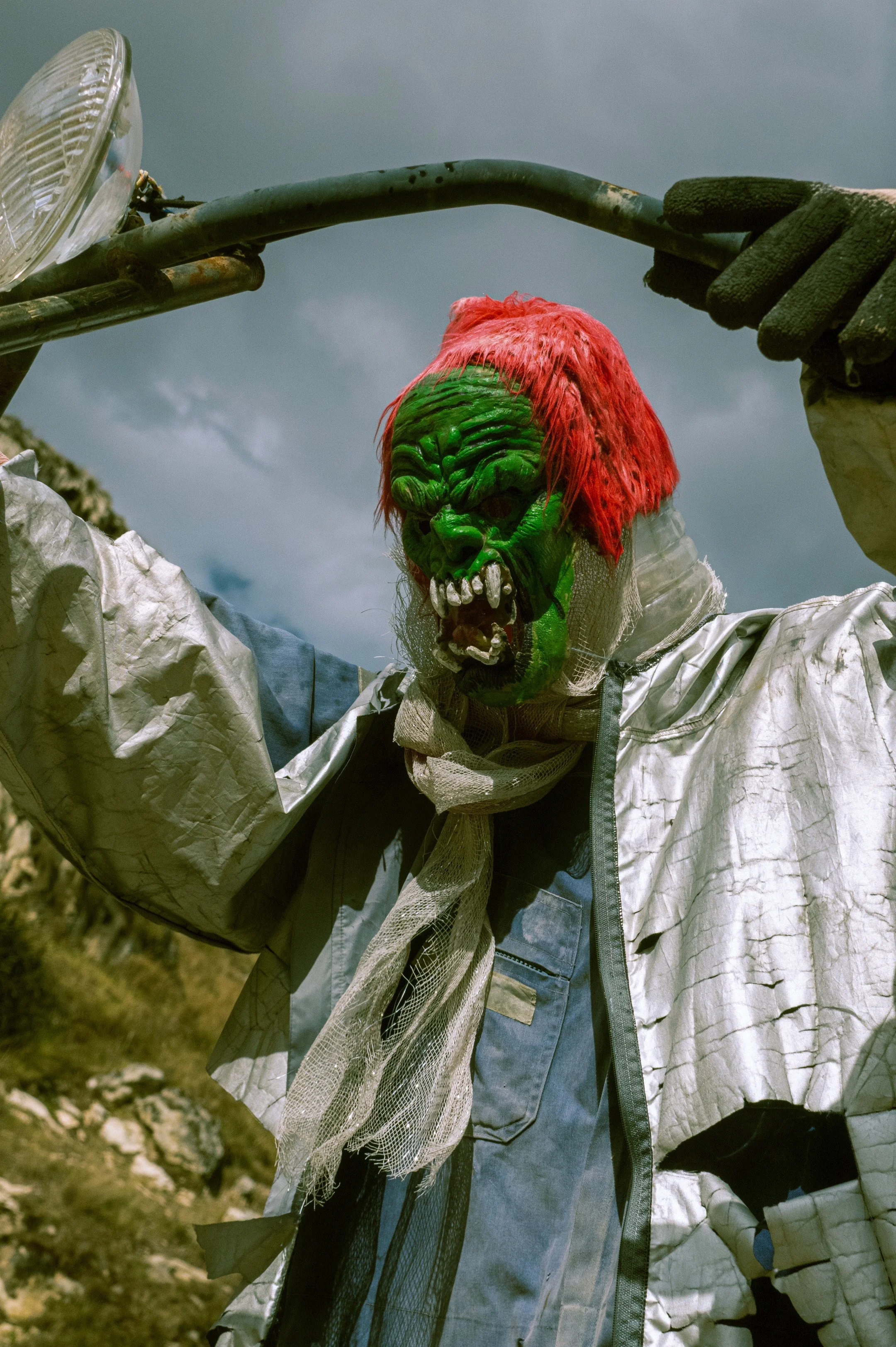 A person wearing a detailed, frightening Halloween mask with green skin, red hair, and sharp teeth, holding a large metal fork against a cloudy sky.