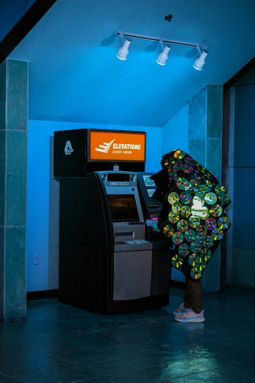 A person in a colorful, disc-covered jacket using an ATM or self-service kiosk in a dimly lit space with blue lighting.