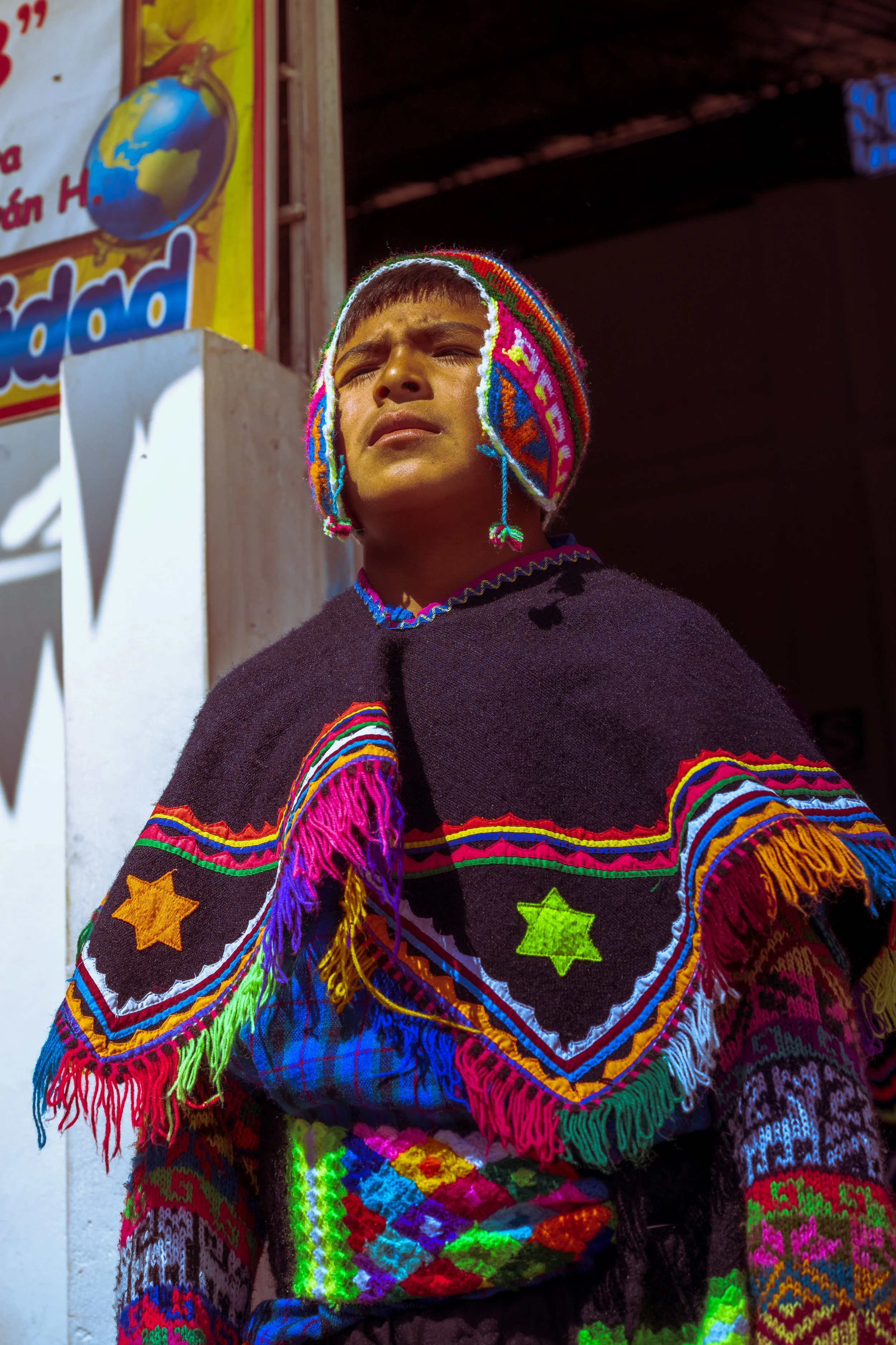 A young boy wearing traditional, colorful, embroidered clothing and a knit hat with bright patterns, standing outdoors near a wall with a sign.
