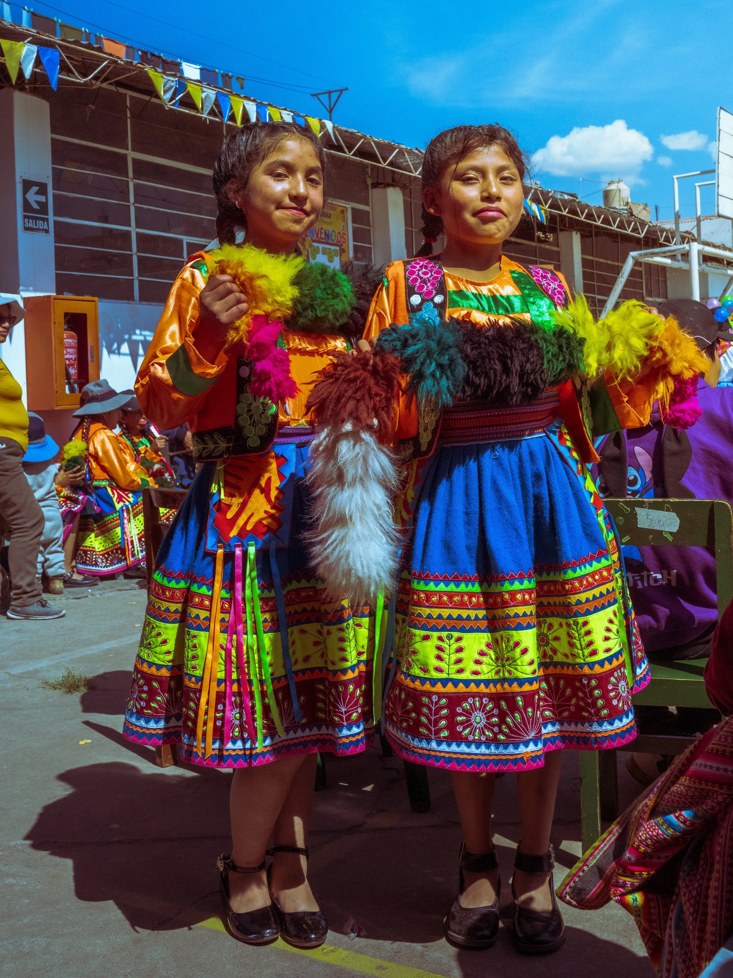Two young women dressed in colorful traditional clothing with intricate patterns in orange, blue, pink, green, and purple, standing outdoors during a cultural celebration, with other people and a building decorated with banners and balloons in the ba