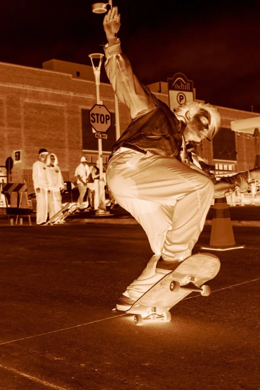 A skateboarder performs a trick on a skateboard at night, with a group of people and street signs visible in the background.