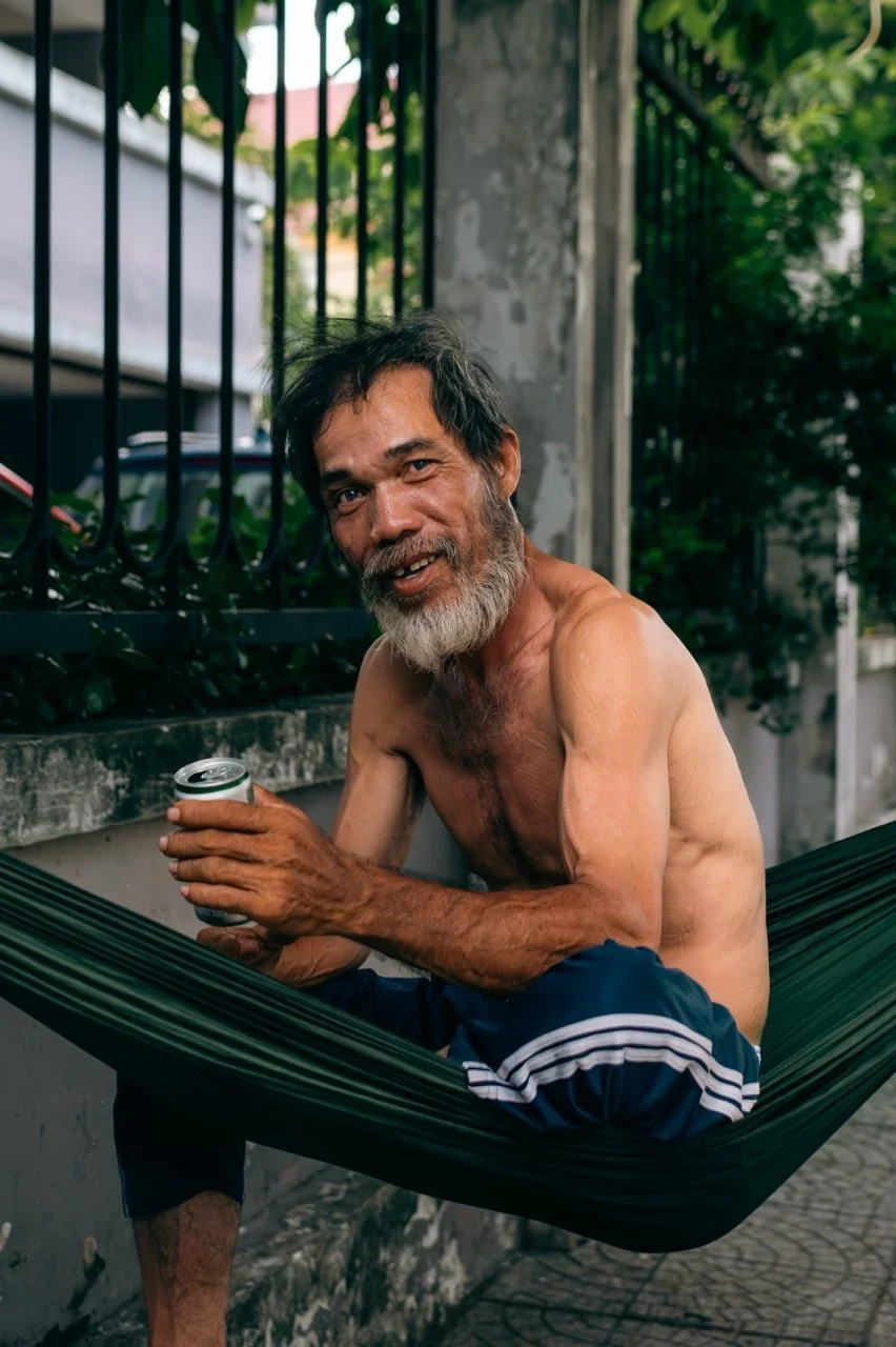 An elderly man with gray hair and beard, shirtless, sitting in a hammock outdoors, holding a can, smiling at the camera.