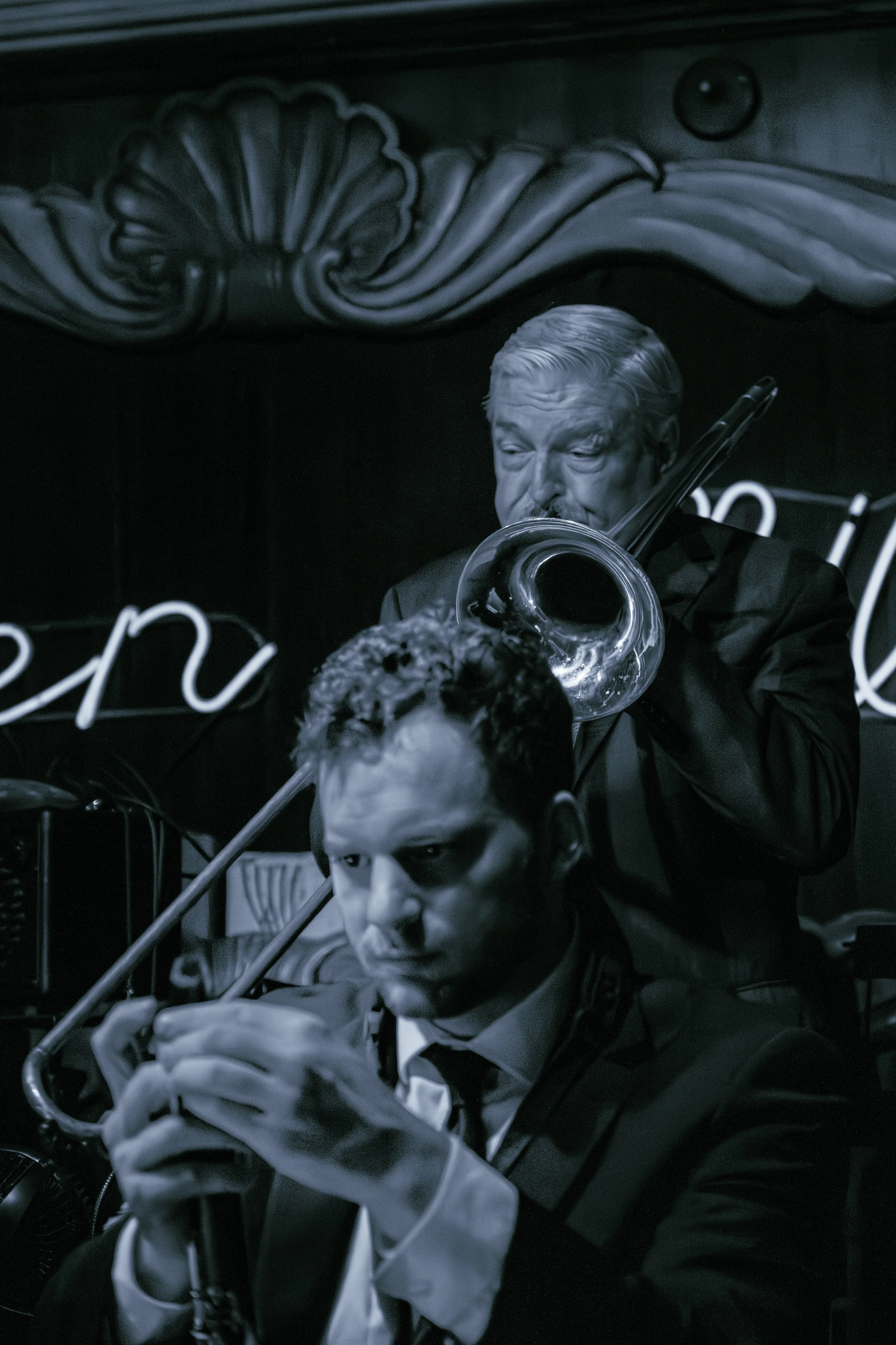 Black and white photo of a man with slicked-back hair playing a trombone, with a woman in front of him also holding a trombone, in a dimly lit setting with a decorated wall and neon sign.
