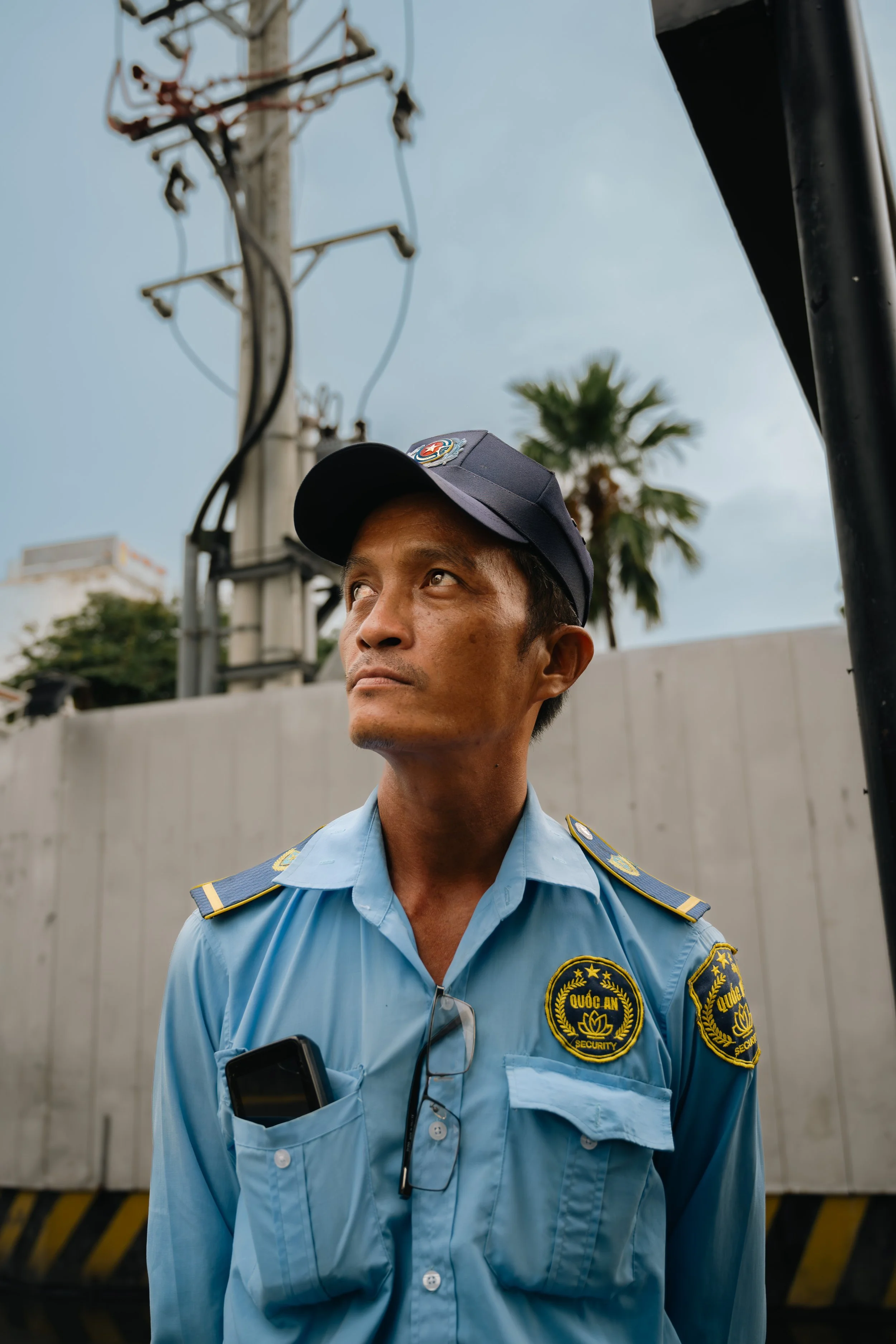 A security guard wearing a light blue uniform and a navy cap stands outdoors near a barrier with a white wall behind him. There are power lines and a palm tree in the background.