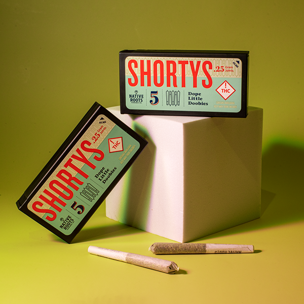 Two boxes labeled 'SHORTYS' with THC and cannabis graphics, one resting on a white cube and one leaning against it, with two pre-rolled joints in front on a yellow-green background.