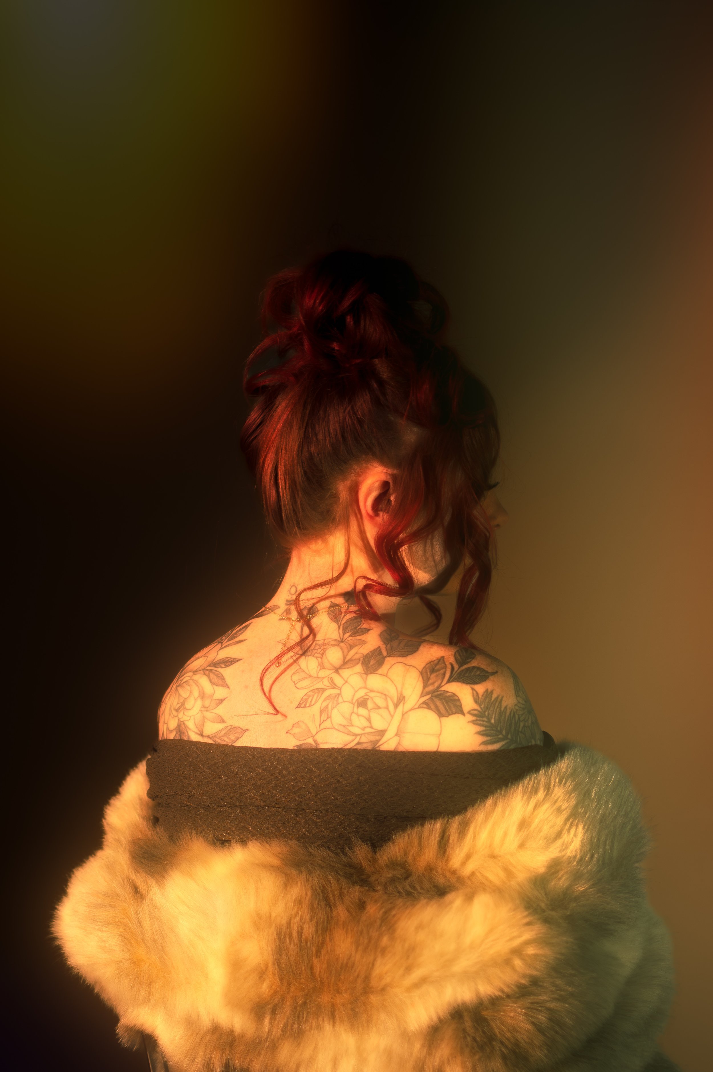 A person with red, curly hair and floral tattoos on the back, wearing a black off-the-shoulder top, sitting on a furry beige blanket with a black background and soft lighting.