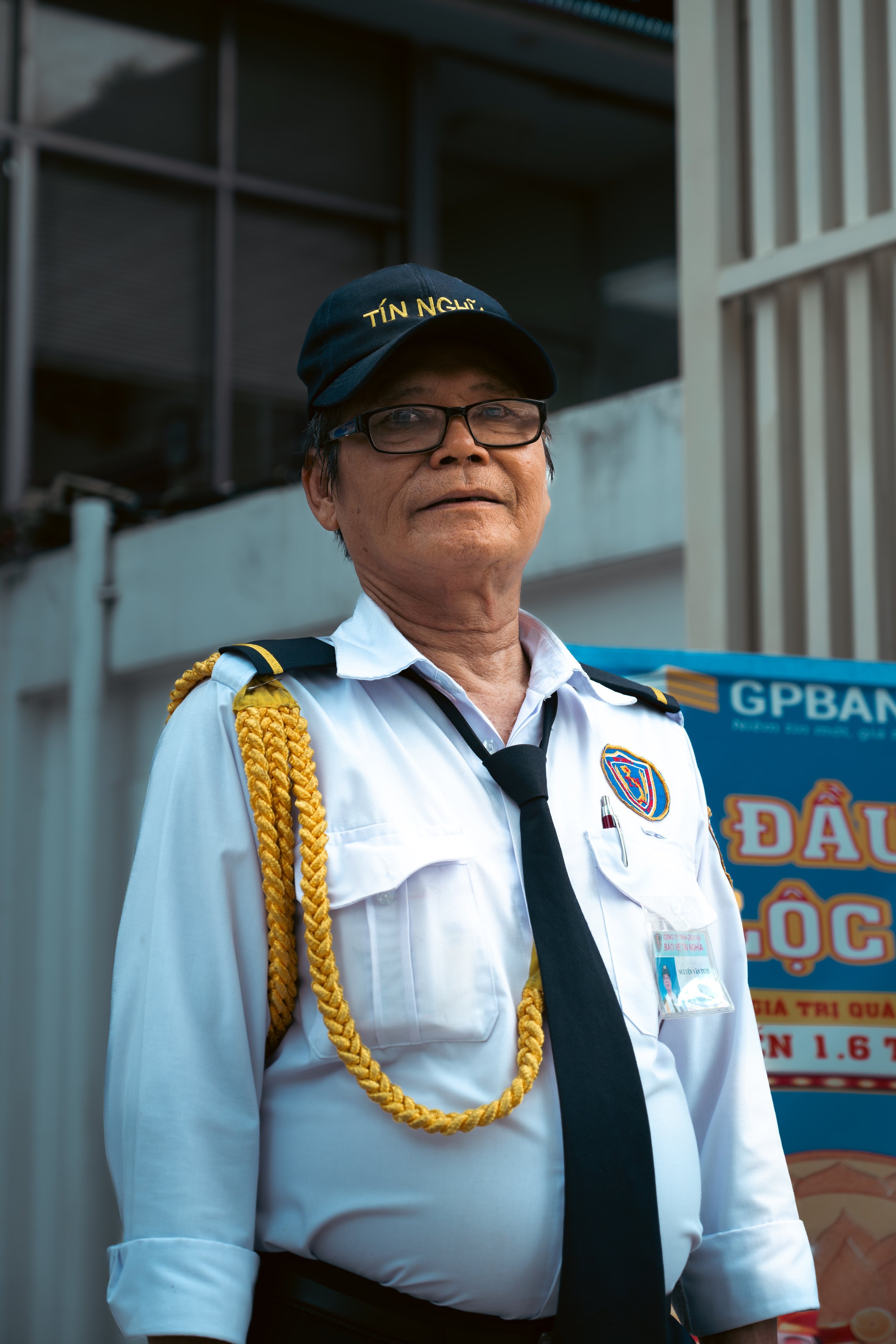 A man wearing a white uniform with a tie and a police badge, a black cap embroidered with yellow text, eyeglasses, and a yellow braided shoulder cord.