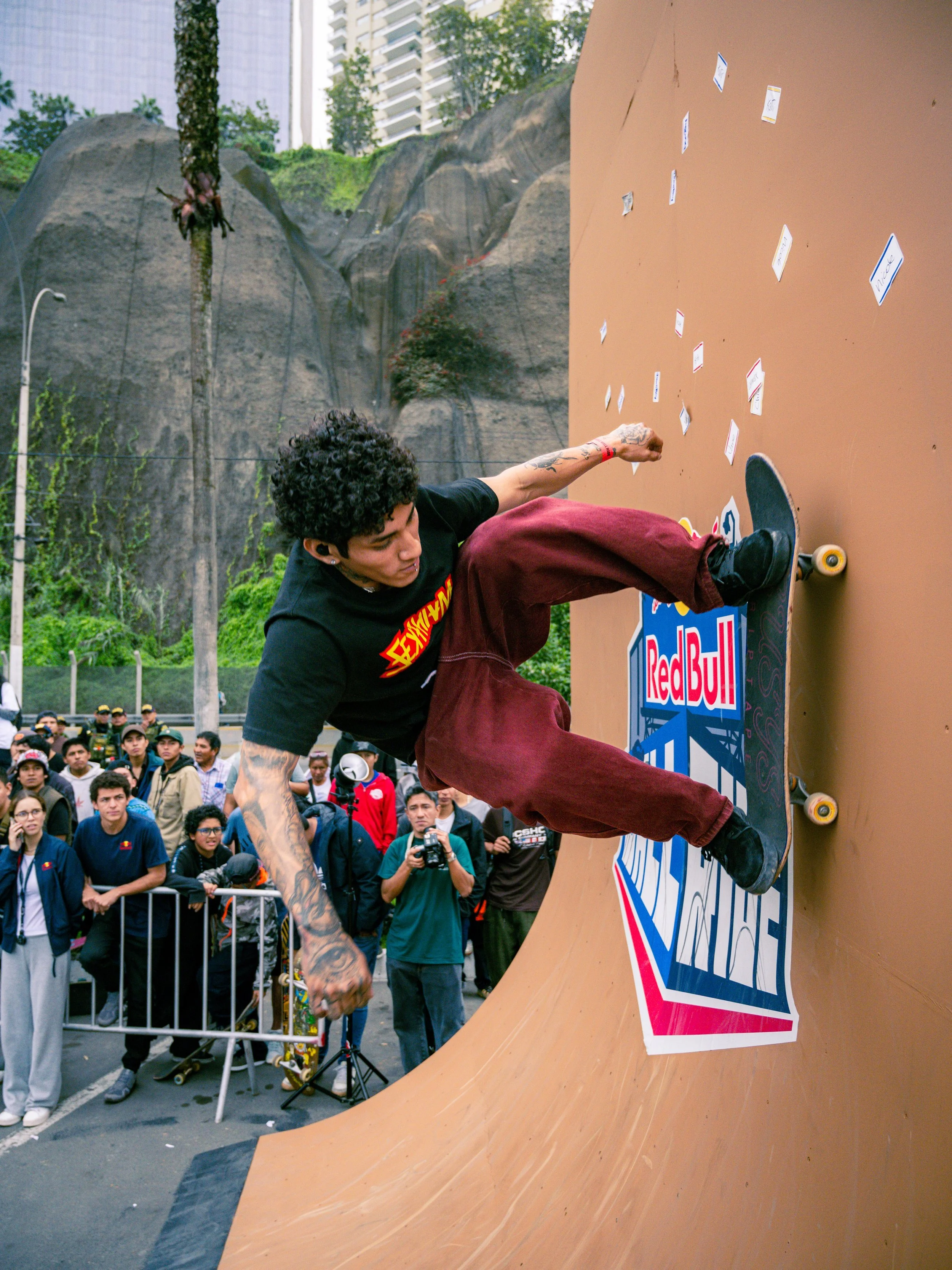 A skateboarder performing a trick on a vert ramp at an outdoor event with a crowd of spectators watching and a cityscape background.