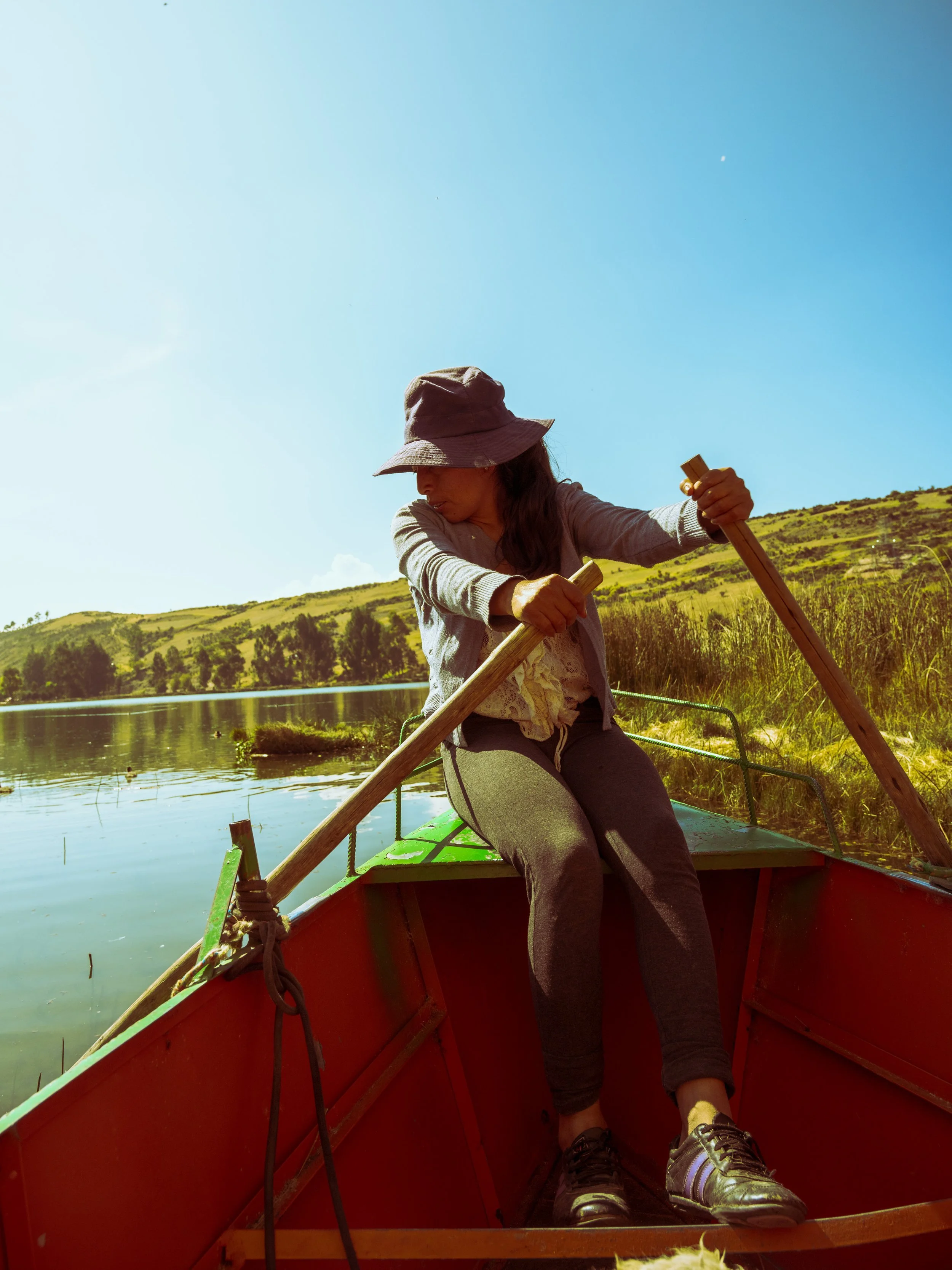 A woman wearing a wide-brim hat and casual clothes is sitting in a red boat on a lake and paddling with a wooden oar. She is near a grassy shoreline with trees and hills in the background, under a clear blue sky.
