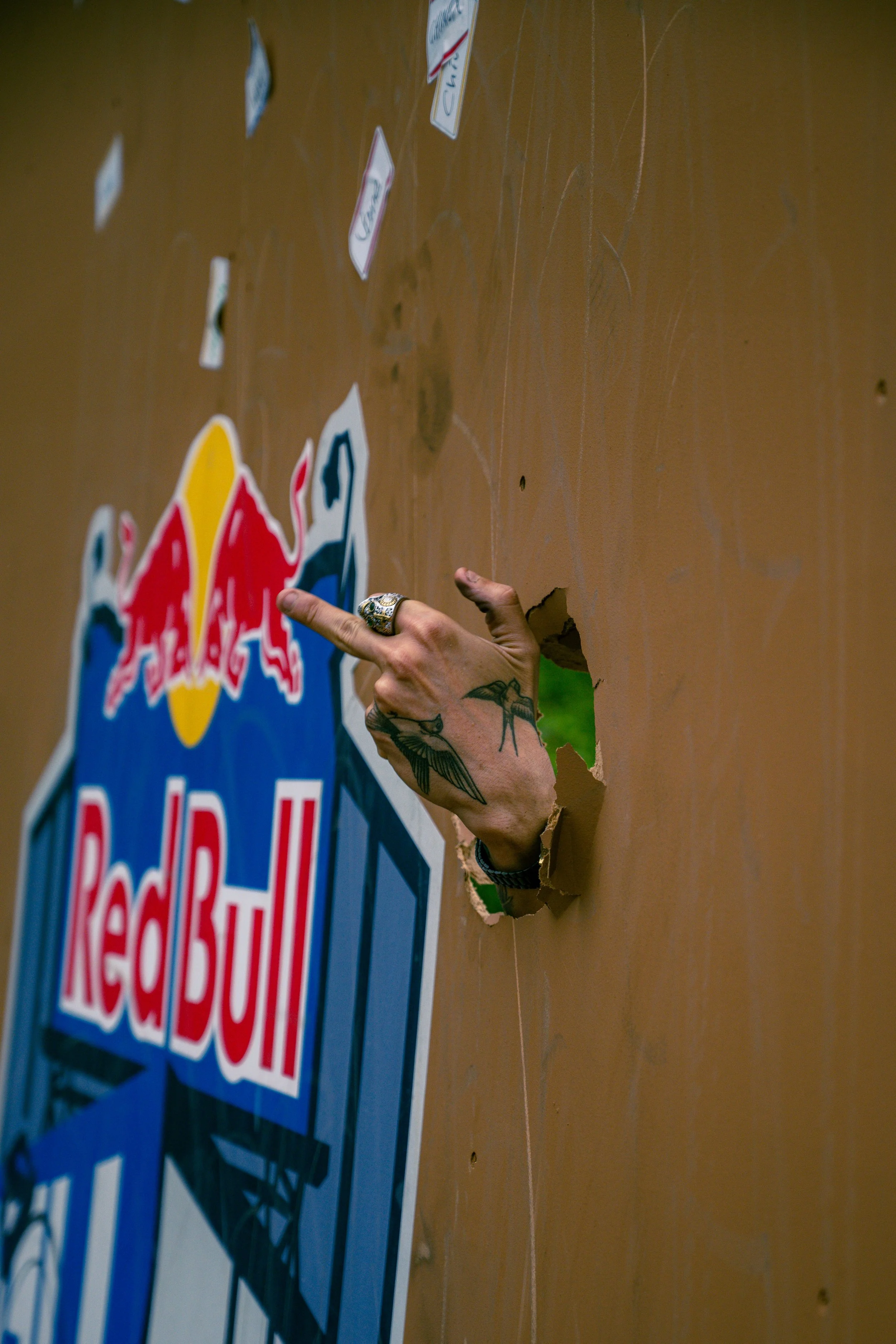 A hand with tattoos and a ring is breaking through a cardboard cutout of a Red Bull logo with a hole in the shape of a hand, against a wooden surface with stickers.