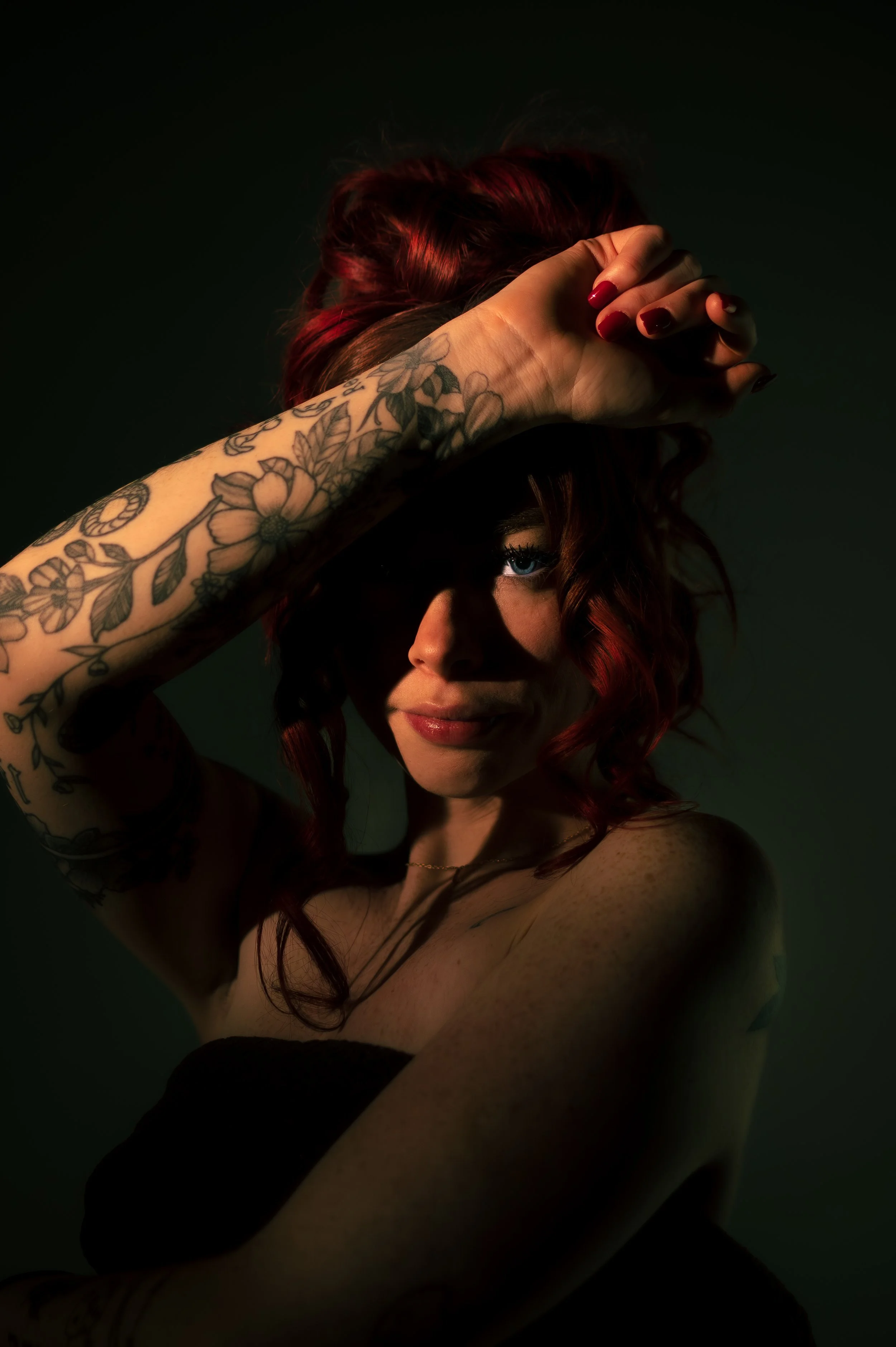 A woman with red hair and tattoos on her arm, covering her face with her hand, revealing one eye, against a dark background.