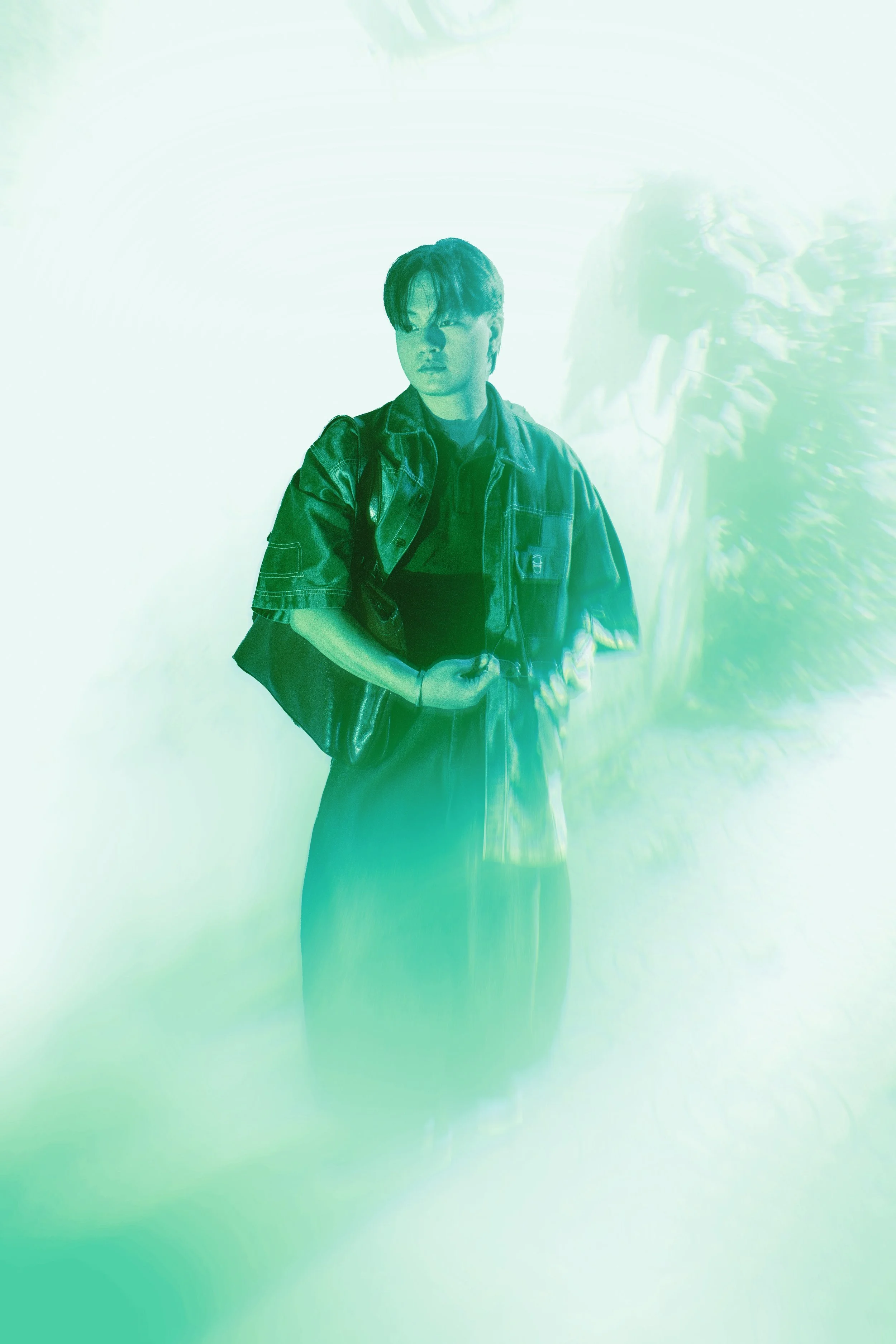 A person with short hair, wearing a dark jacket and carrying a backpack, stands against a wall with colorful, abstract light projections.