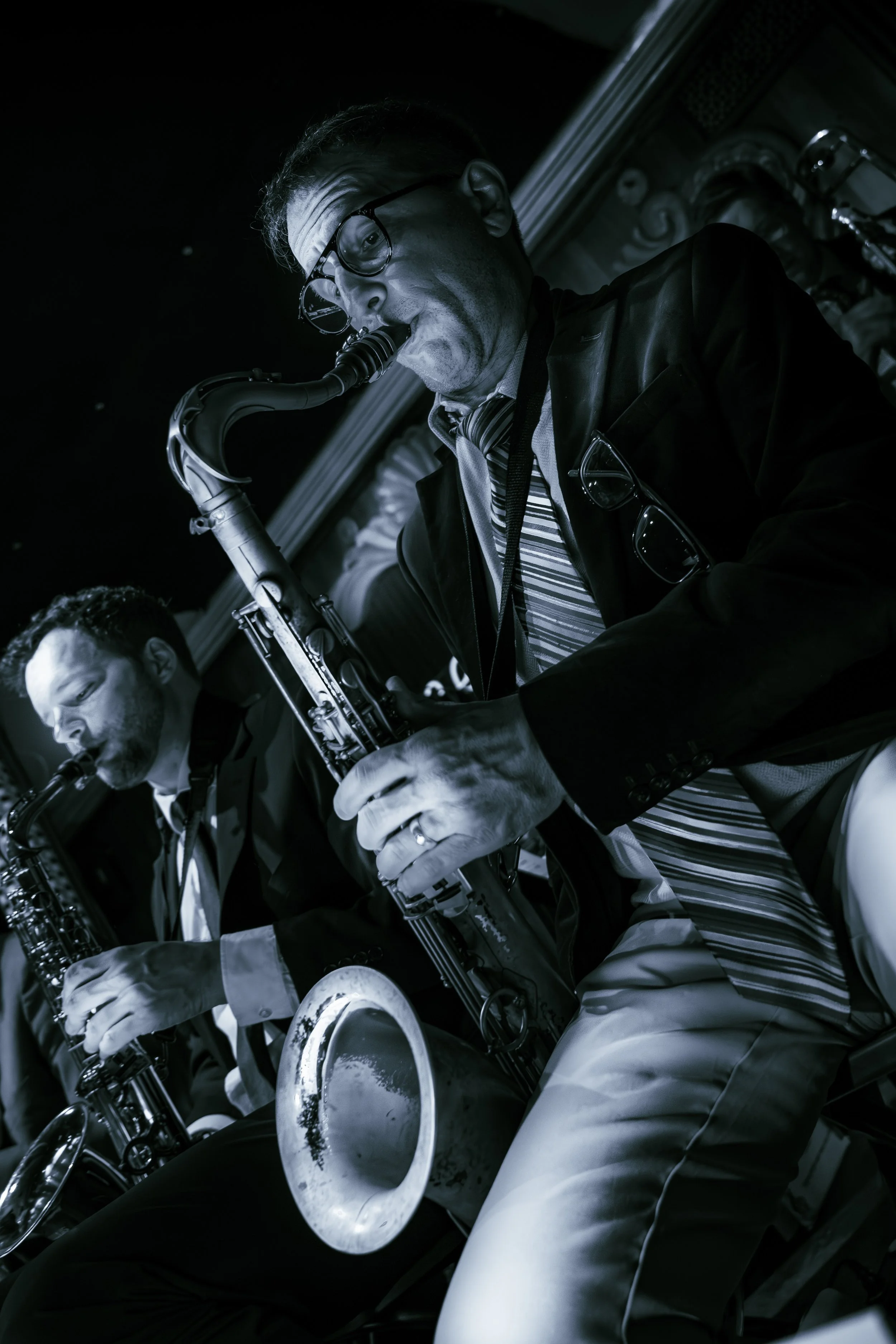 Black and white photo of two men playing saxophones. The man in the foreground is wearing glasses, a striped shirt, and a dark jacket, with a pair of glasses hanging from his collar. The other man in the background is dressed in a light-colored shirt