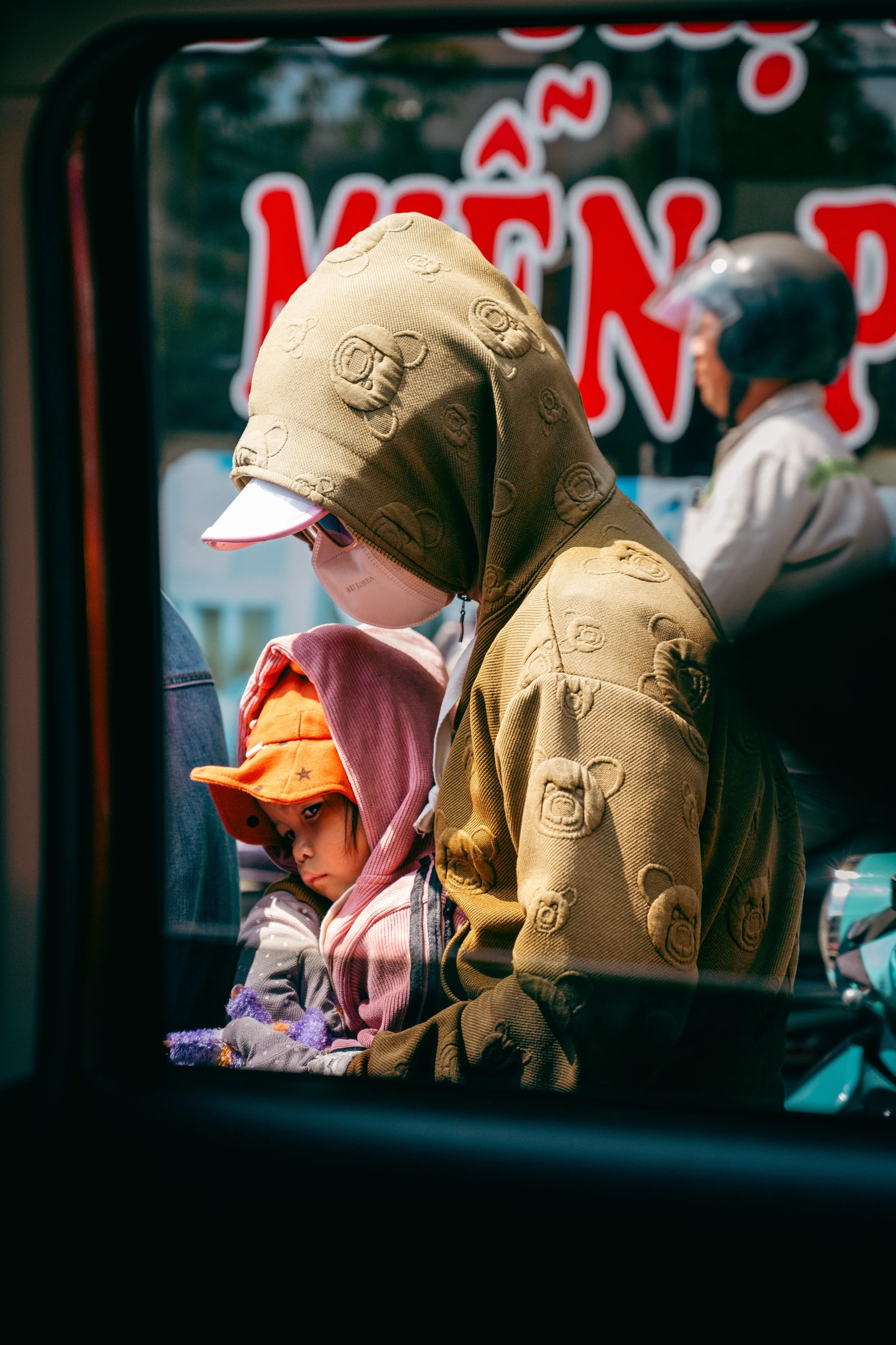 People wearing face masks and hoodies are seen through the window of a moving vehicle, with a red and white sign in the background.