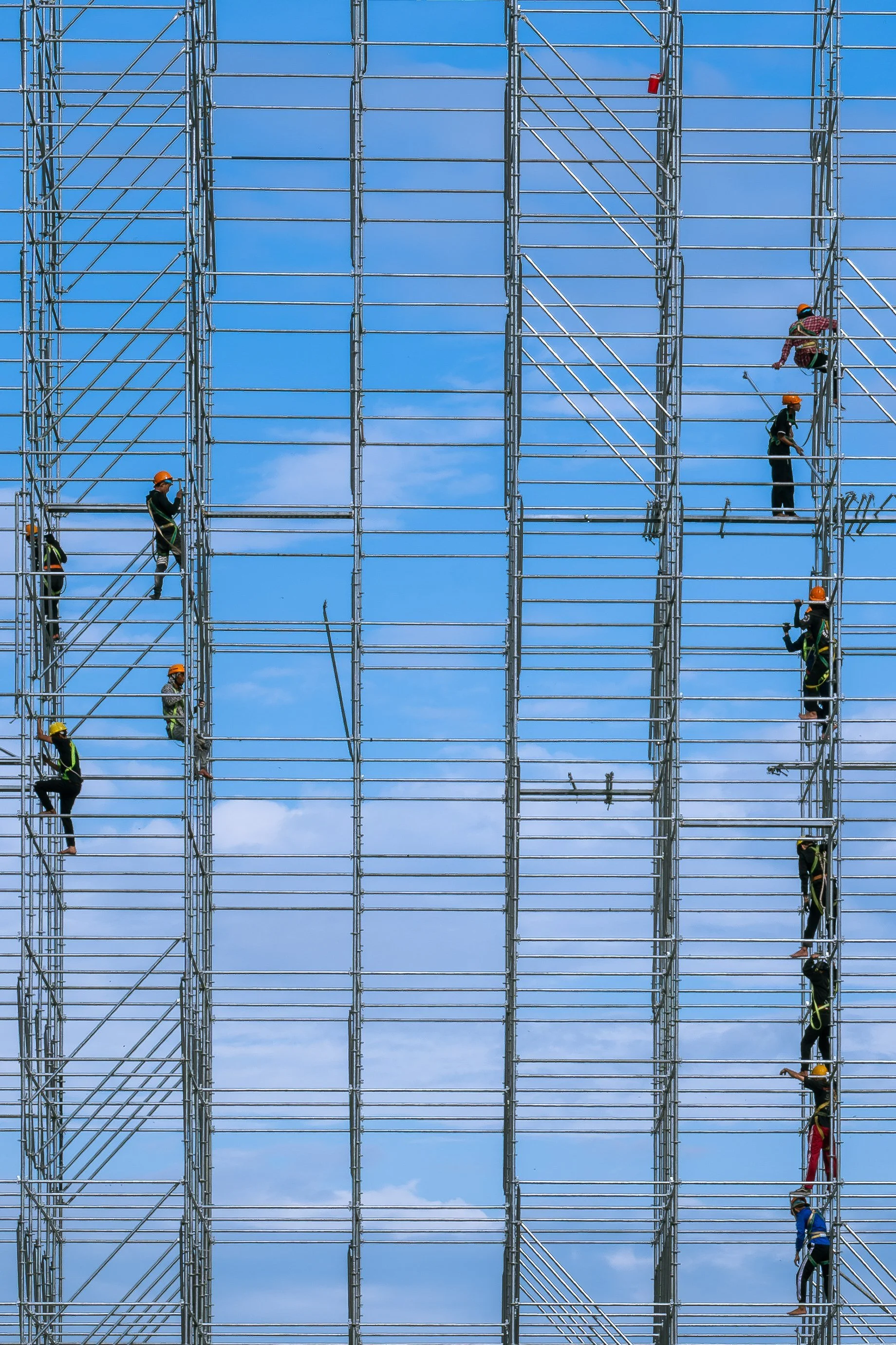 Construction workers wearing safety helmets climbing and working on a large metal scaffolding structure against a background of a clear blue sky.